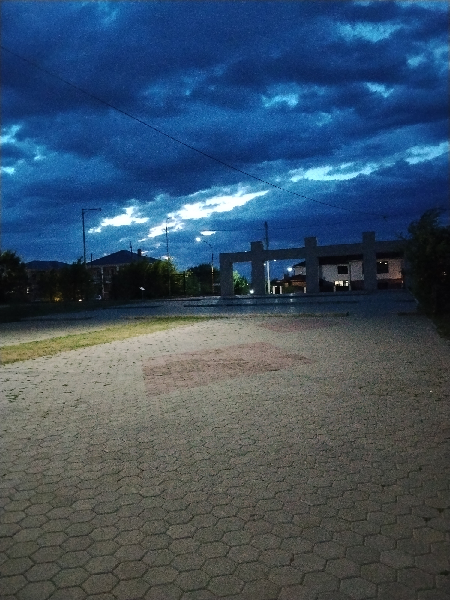The image depicts an outdoor scene at dusk, likely in Kokshetau, Kazakhstan, given the context. The sky is dominated by dramatic, dark blue clouds, with streaks of white light breaking through, suggesting the last moments of daylight or impending weather. In the foreground, a hexagonal-patterned paved area extends into the distance. A patch of grass with scattered reddish-brown stones is visible to the left of the paving.
Further back, a modern architectural structure with four large, concrete pillars supporting horizontal beams stands in the center. Behind this structure, a light-colored building with dark windows is partially visible. The building appears to be illuminated from within. Trees and other vegetation are present on either side of the building and the concrete structure. Streetlights, one tall and slender on the left and another with a spherical lamp on the right, are also visible, casting a faint light. A power line is strung across the upper portion of the sky. The overall impression is a public space, possibly a monument or a plaza, under a moody evening sky. There are no people or animals visible in the image.