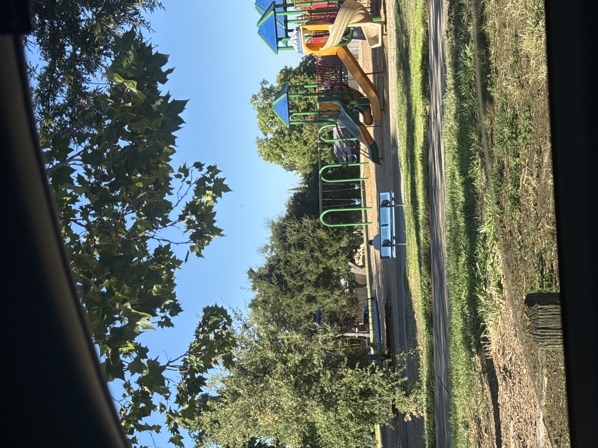 The image shows a playground in a park. In the foreground, there is a patch of grass and a weathered wooden post. Beyond that, there is a paved pathway separating the grassy area from a wood chip-covered section of the playground. In the midground, there is a blue bench with a person sitting on it. To the right of the bench, there is a colorful playground structure with slides and swings. The playground is surrounded by trees and lush green foliage. The sky is a clear blue. The overall scene depicts a sunny day at a recreational park.
