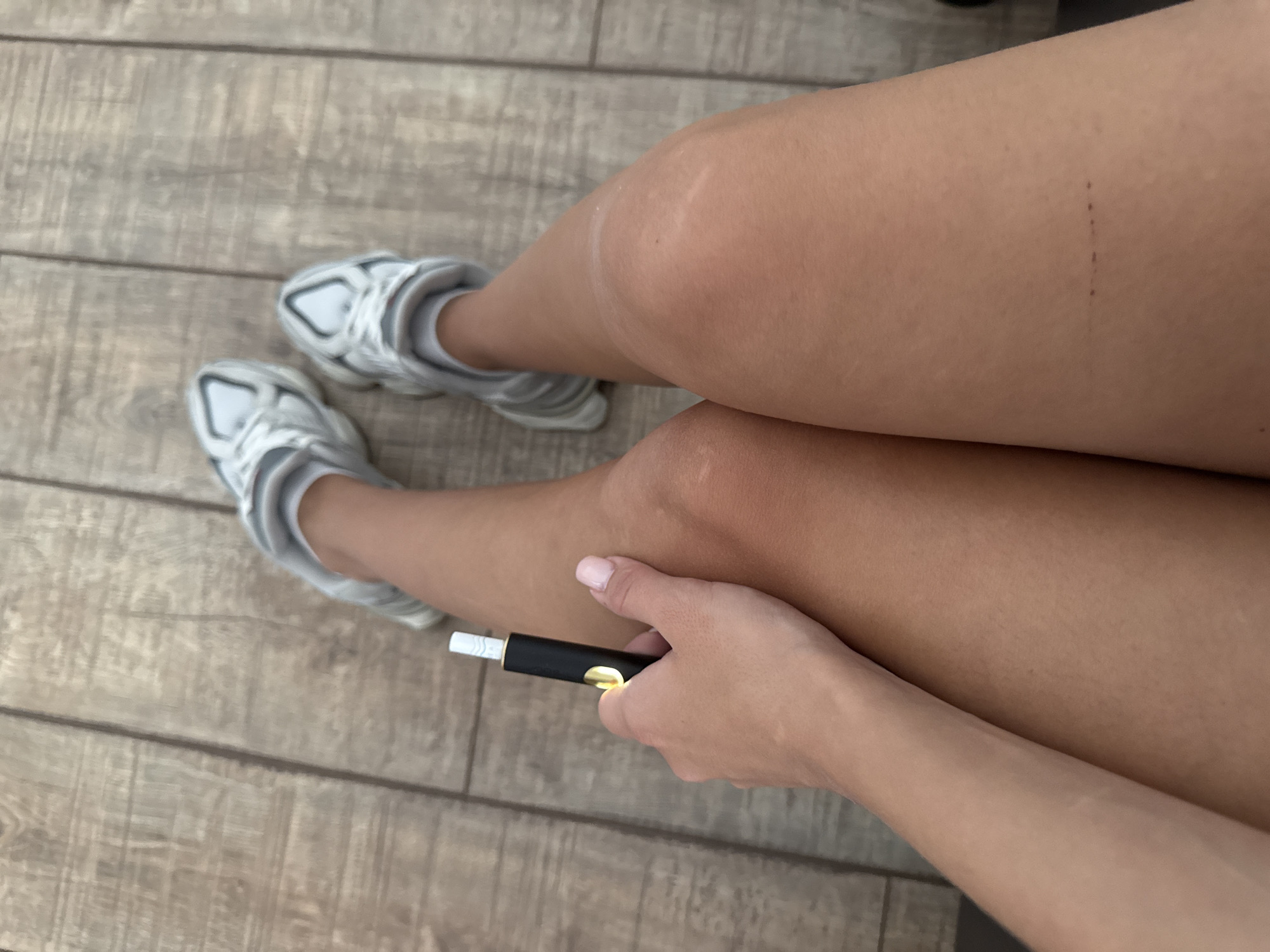 A close-up, overhead view shows a person's legs and feet, clad in white athletic shoes and socks, resting on a wooden floor. In the foreground, the person's left hand, with lightly manicured nails, is holding a black and gold electronic cigarette device. The scene appears to be indoors, on a wooden floor that might be found in a home, a business, or a public space within Chișinău, Moldova. There are no other people or significant environmental details visible.