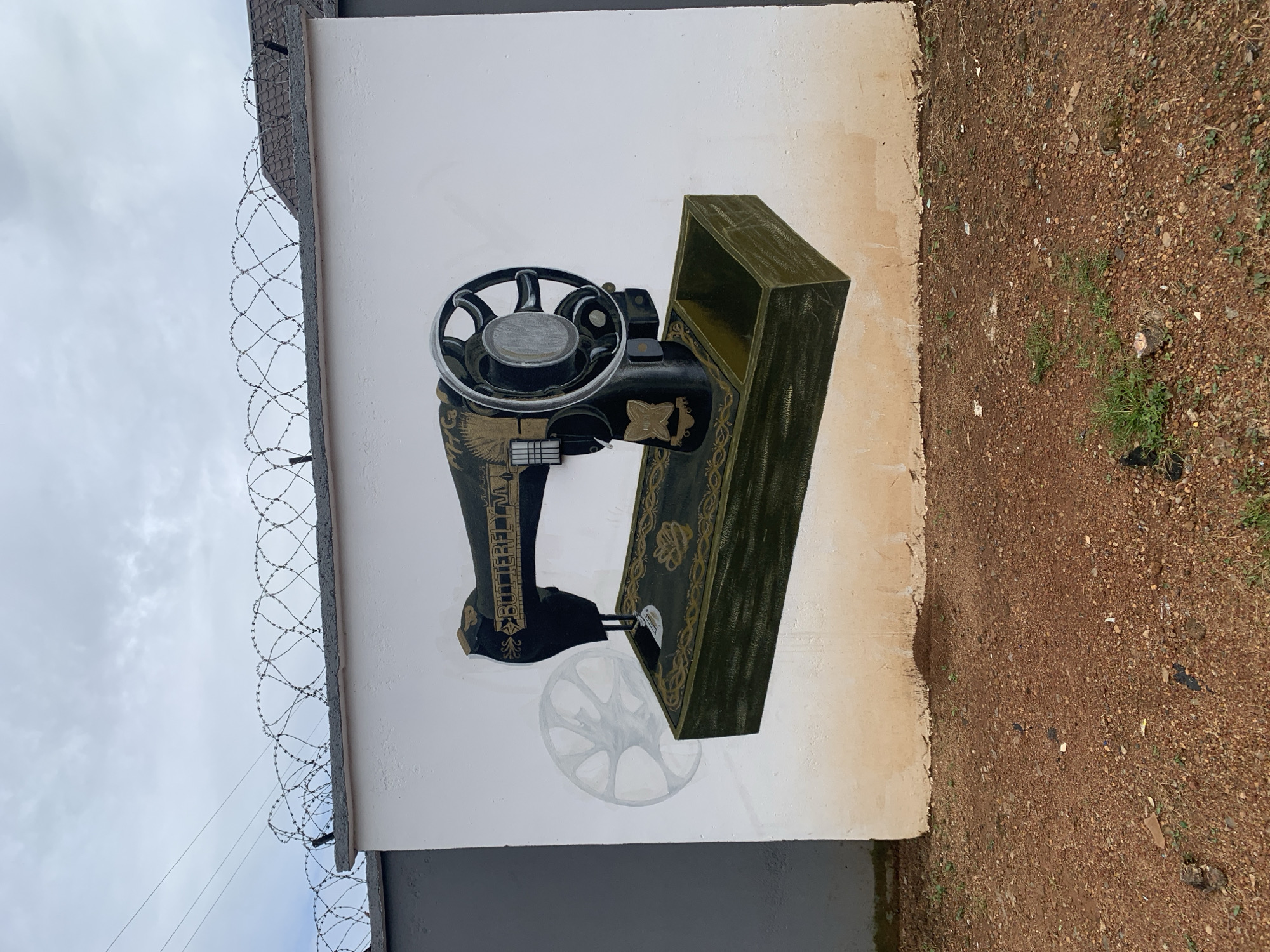 In Gura Top, Nigeria, a mural of a vintage sewing machine is painted on a white concrete wall. The sewing machine is black and gold, with the brand name "BUTTERFLY" visible on the arm. It sits on a green base with ornate gold detailing. To the left of the machine, a faded, ghost-like image of a spinning wheel is painted on the wall, suggesting movement. The wall itself is bordered at the top by a tangle of barbed wire, indicating a security measure. The ground in front of the wall is covered in reddish-brown gravel and sparse green vegetation. The sky above is overcast.