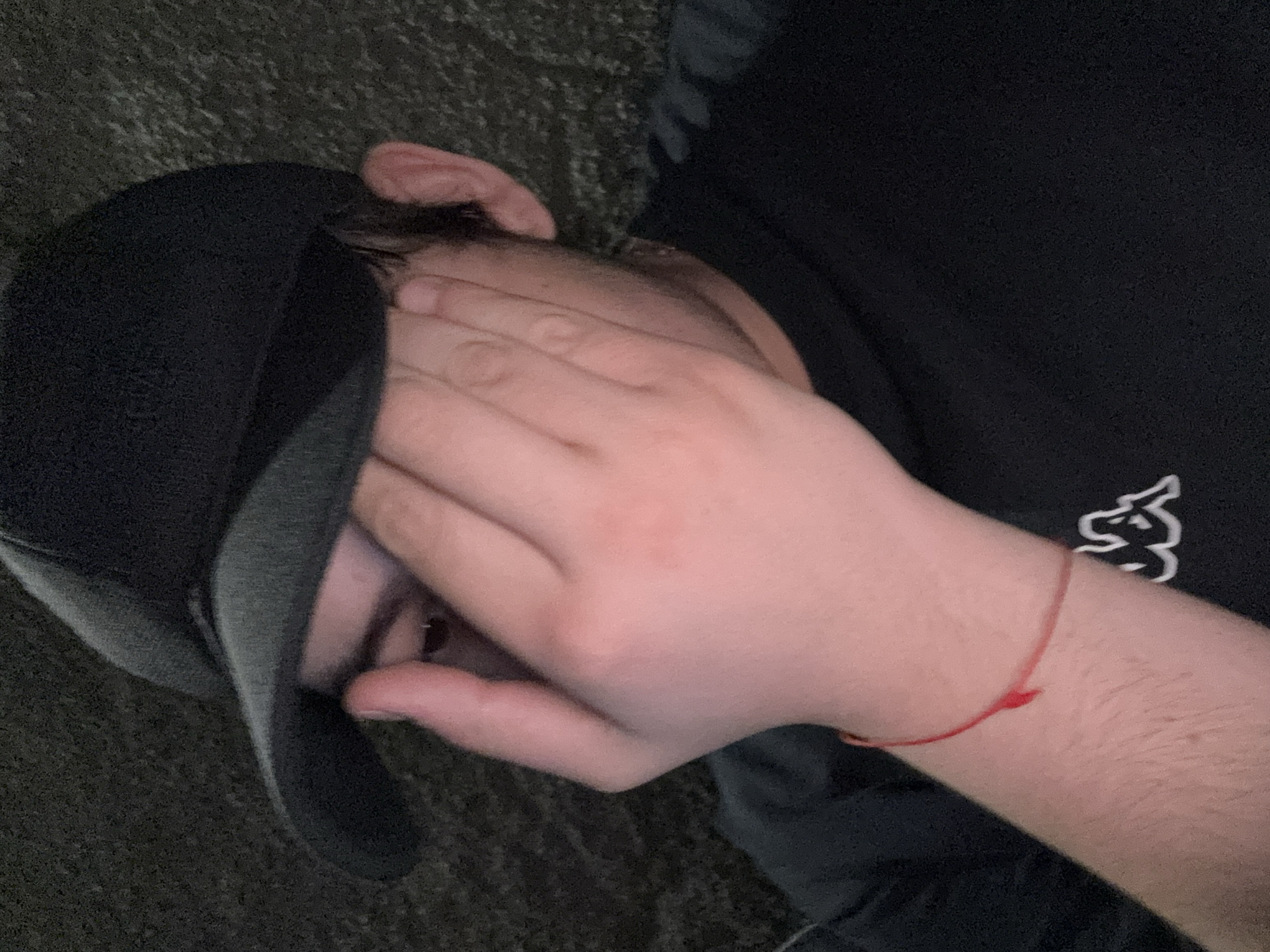 The image shows a person lying on what appears to be a dark, textured surface, possibly concrete or asphalt. The person is wearing a black t-shirt with a white logo visible on the upper right. They have a black baseball cap on their head, tilted downward. Their face is mostly obscured by their hand. A red string bracelet is visible on their wrist. The location, Yerevan, Armenia is not discernible from the image.