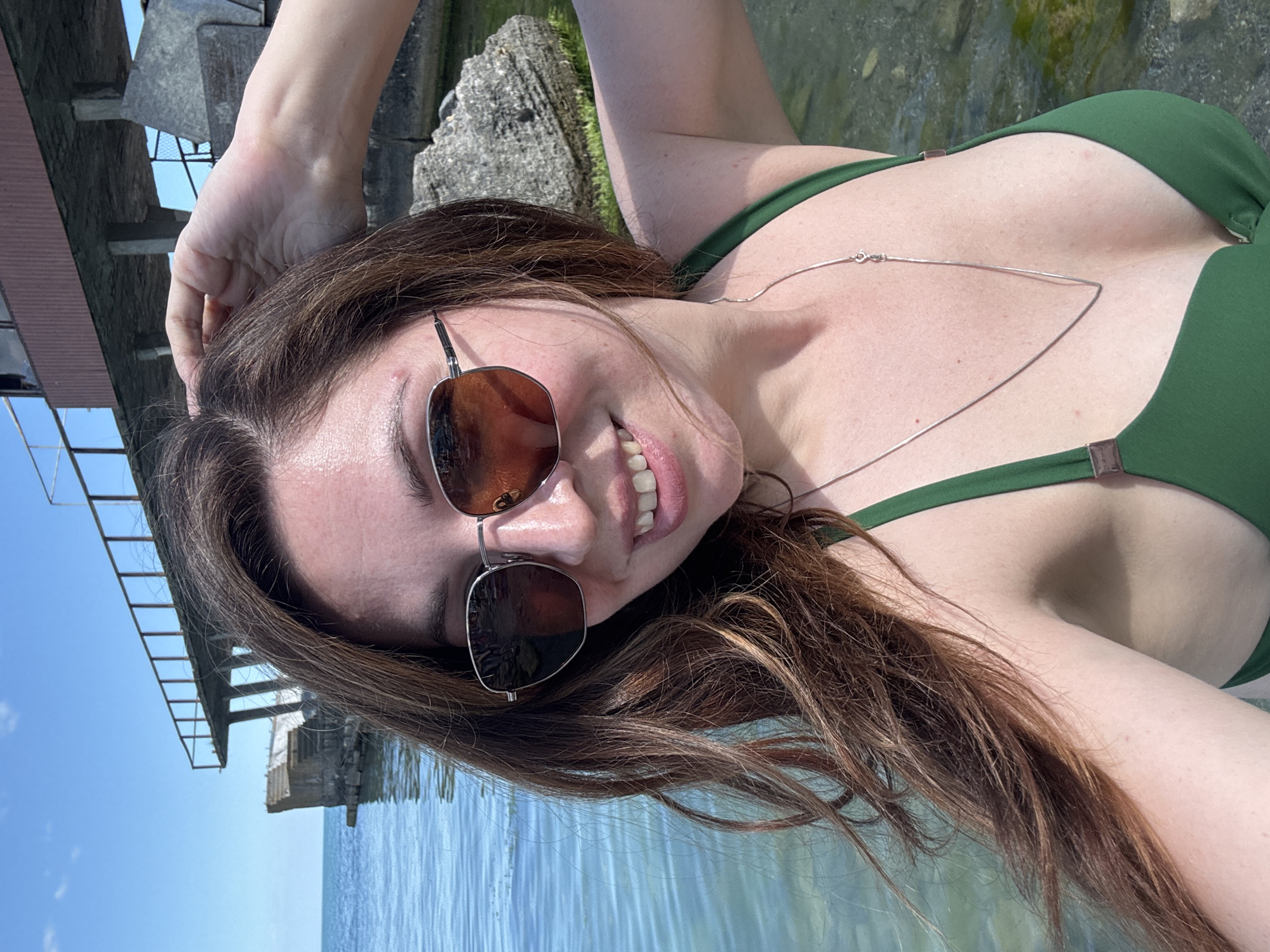 A woman with long, brown hair and sunglasses is taking a selfie in the water near some concrete structures. She is wearing a green bikini and a silver necklace. The water is clear and blue, and the sky is also blue with some clouds. In the background, there is a large concrete structure with railings, possibly a pier or a platform. There are also rocks and vegetation along the water's edge. The scene appears to be a sunny day at a seaside location in Odesa, Ukraine.