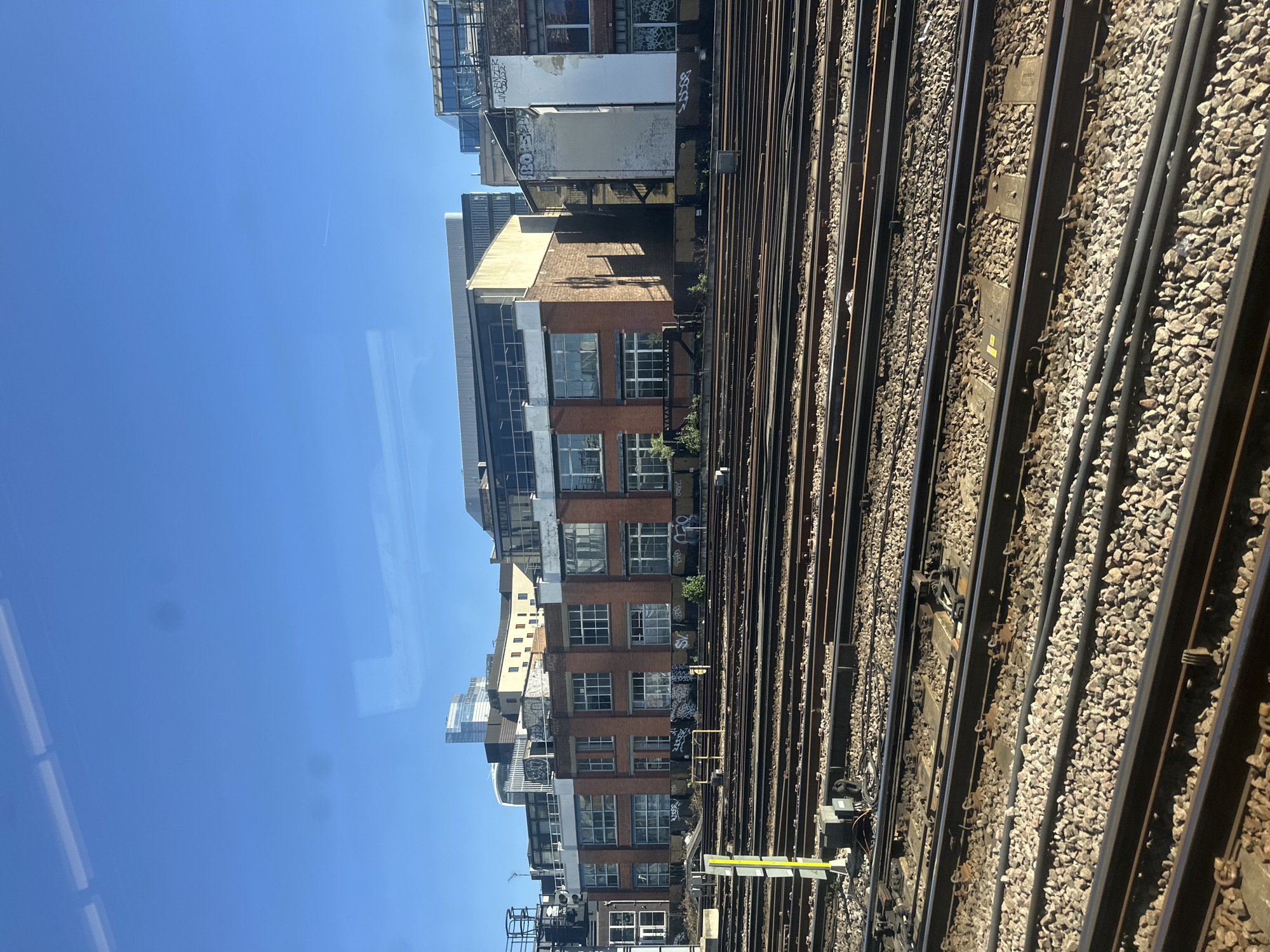 This media file presents an urban landscape, likely captured from an elevated perspective such as a train or platform window in London, United Kingdom. The image is oriented vertically but depicts a scene that would typically be viewed horizontally, with a clear blue sky dominating the left side, and railway tracks and buildings on the right.
The upper-left portion of the image shows a vast, clear blue sky, indicating bright daylight. Faint reflections, possibly from internal lights or structures of the viewing platform/train, are visible on the sky's surface, along with a barely discernible contrail from an aircraft high above.
The right side of the image is filled with an intricate network of parallel railway tracks, set upon light grey gravel ballast. Dark metal rails stretch into the distance, accompanied by various black cables or conduits running alongside. Several yellow-and-black trackside markers or small signal posts are scattered among the tracks; the text on these markers is too small to be legible, but they signify track infrastructure.
Beyond the railway lines, a dense row of multi-story urban buildings forms the backdrop. These structures exhibit a variety of architectural styles, ranging from older red-brick buildings with numerous gridded windows to more modern constructions featuring sleek glass facades and lighter-colored materials. Many of the buildings, particularly the older brick ones, are adorned with extensive graffiti and tags, including visible markings such as "BOBO" and "SOWA" (or "SOFA"). The mix of weathered, graffitied structures and contemporary glass towers highlights the diverse urban fabric.
The time of day is clearly bright and sunny, evidenced by the clear sky and distinct shadows cast by the buildings and trackside elements. No people are visible in the scene. The overall impression is one of a bustling urban environment viewed from a transit corridor.