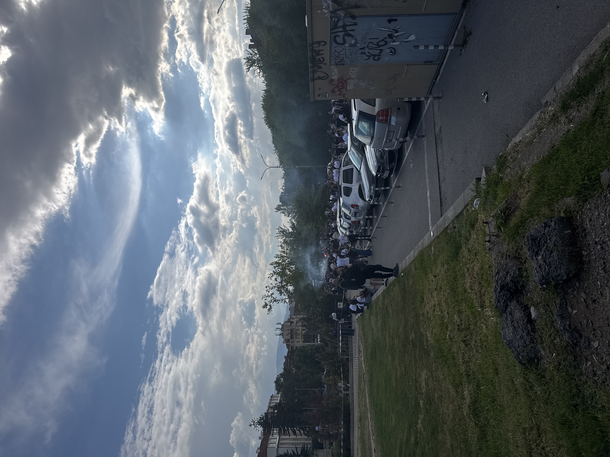 The image depicts a crowd of people gathered along a street in Sofia, Bulgaria. The crowd is largely composed of individuals wearing white shirts. Smoke is visible rising from within the crowd, suggesting the use of flares or pyrotechnics. Several cars are parked along the side of the street. A building with graffiti on its side is adjacent to the parked cars. Across the street, there's a grassy area with trees and buildings in the background. The sky is partly cloudy with sunlight breaking through.