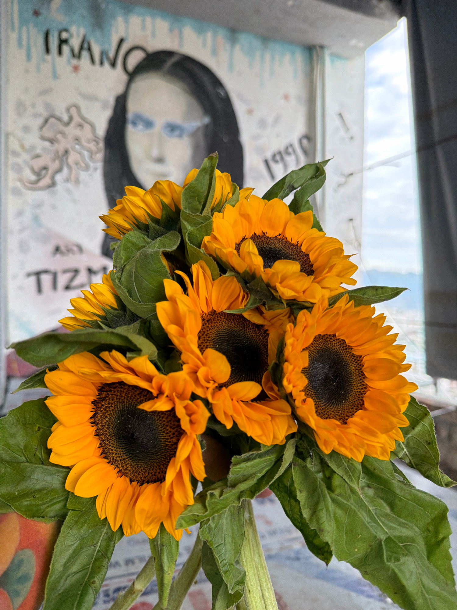 In Nha Trang, Vietnam, a bouquet of sunflowers sits in the foreground, their bright orange petals and dark centers contrasting with lush green leaves. Behind the flowers, a painted mural is visible on a wall. The mural features a portrait of a person with dark hair and striking blue eyes, partially obscured by the flowers. Text elements, including "IRAN" and "1990," are also present on the mural, along with other artistic designs. To the right of the mural, a window reveals a cloudy sky and a distant view of buildings or mountains. The overall scene appears to be an indoor setting with an outdoor view, possibly on a balcony or near a window.