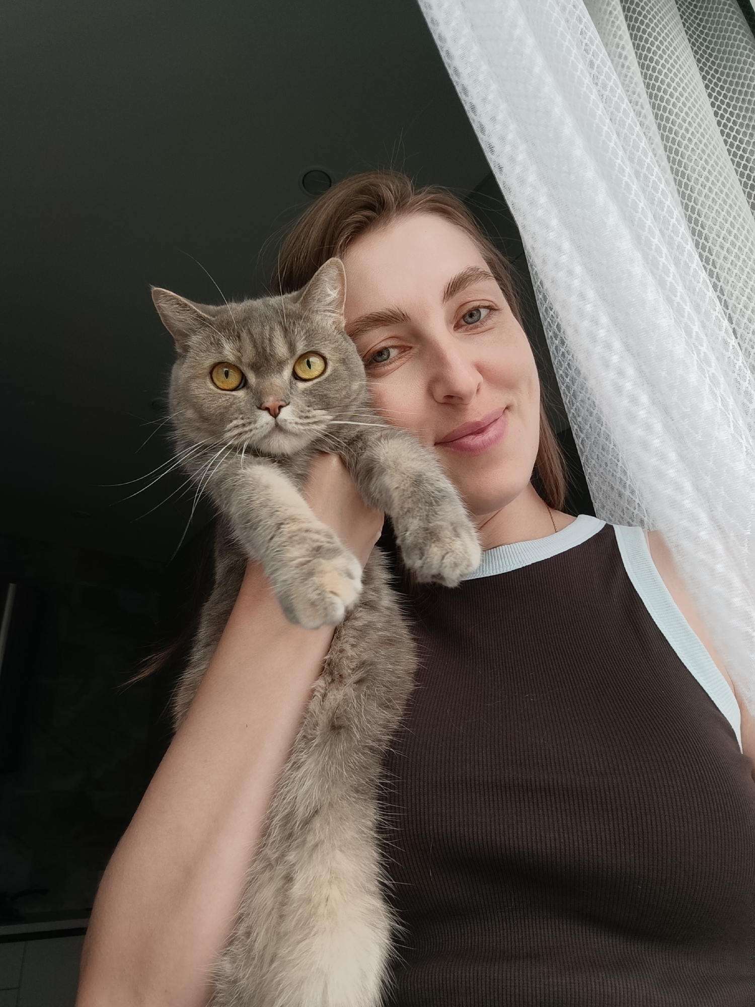 A woman is holding a grey tabby cat while posing for a selfie. The woman has fair skin and brown hair, and she is smiling at the camera. The cat, with striking yellow eyes, is nestled in her arms, its paws extending outwards. The background is divided into two parts: on the left, a dark, out-of-focus area suggests an interior space, possibly a room with a window; on the right, a white, lacy curtain is visible, letting in soft light. The overall lighting suggests it might be daytime. The woman is wearing a dark brown tank top with a light blue trim. The scene appears to be set indoors, near a window, with a relaxed and affectionate mood between the woman and her cat. There is no visible text in the image.