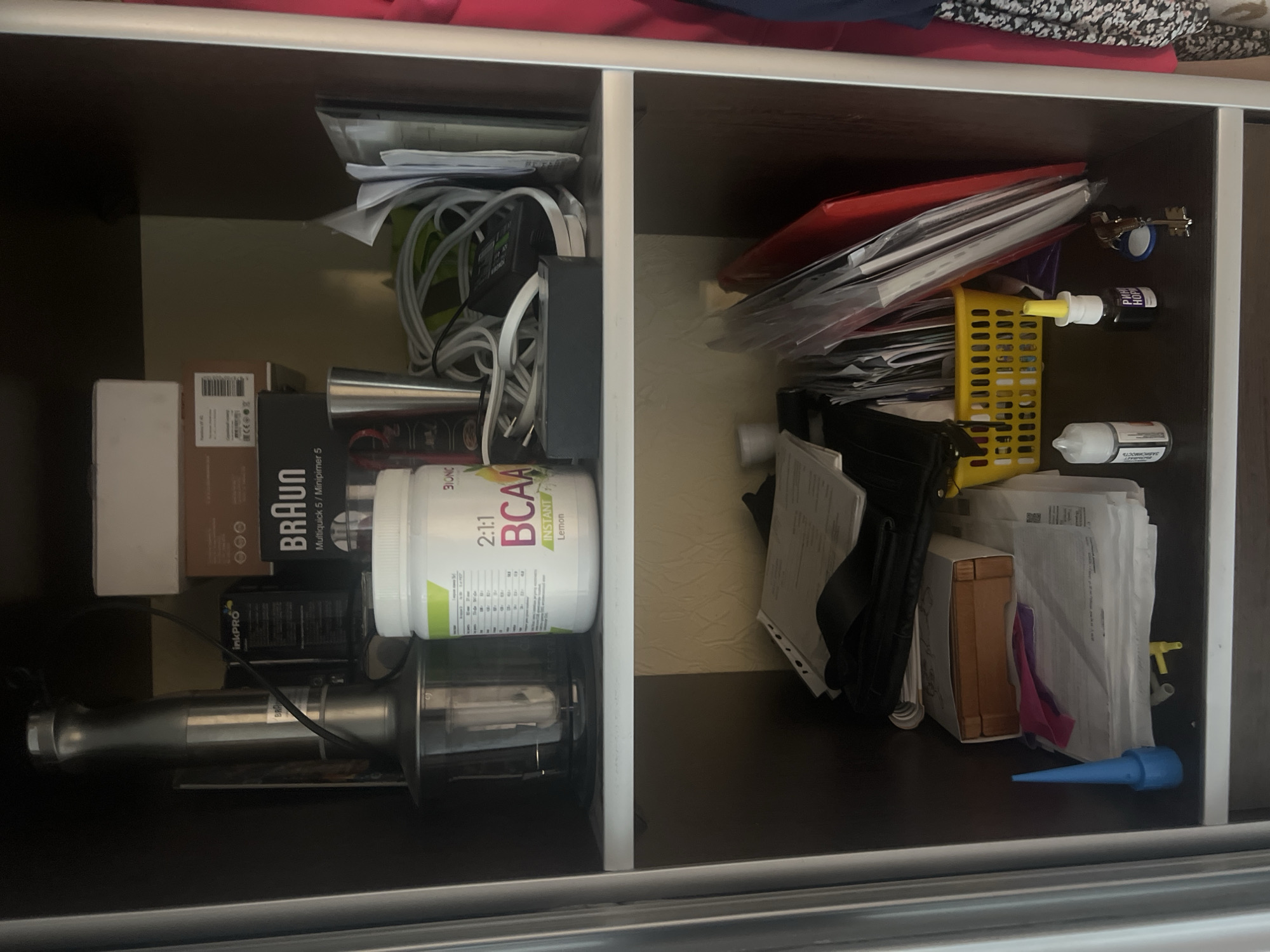 The image shows the interior of a dark brown, two-shelf closet. The top shelf contains a silver immersion blender with its accessories in a plastic container, a white box, a brown box with a black Braun Multiquick 5 label, and a white tub of BCAA supplements. Also on the top shelf are various wires and chargers. The bottom shelf holds stacks of paper, a wooden box, a blue plastic cone-shaped object, a yellow basket, a red folder, and other various small items including bottles and keys. The background of the closet appears to be a textured beige wallpaper. The image is taken from a slightly low angle, looking up into the closet. The location of Veteran-2, Russia, is not discernible from the image itself.