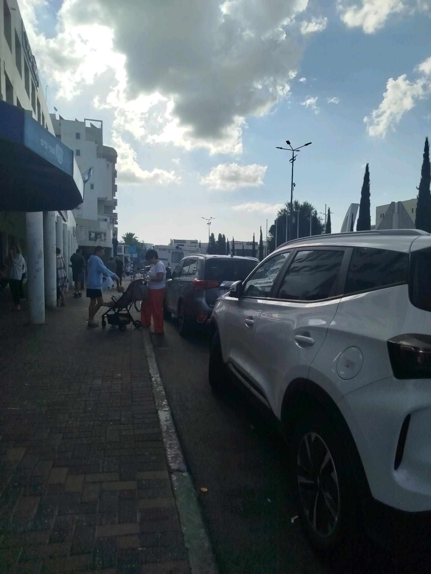 The image shows a street scene in Ashkelon, Israel. In the foreground, a white car is parked on the right side of the road. The car appears to be a modern SUV with tinted windows. To the left of the car is a sidewalk made of brick pavers, which is slightly wet. A light-colored curb separates the sidewalk from the road.
On the sidewalk, several people are visible. A person in a light blue hoodie and dark shorts is pushing a stroller. Another person, wearing orange pants and a white top, is standing next to the stroller. There are other pedestrians in the background, some walking and some standing.
Behind the sidewalk, there is a multi-story building with white facades and dark-tinted windows. The building has a modern architectural style. A dark blue awning or overhang extends from the building over the sidewalk. The sky above is partly cloudy with patches of bright sunlight.
Further in the background, more buildings and structures can be seen, including tall, dark green trees that appear to be cypress trees. A street lamp with multiple lights is also visible. The overall impression is a typical urban street with pedestrian and vehicular traffic.