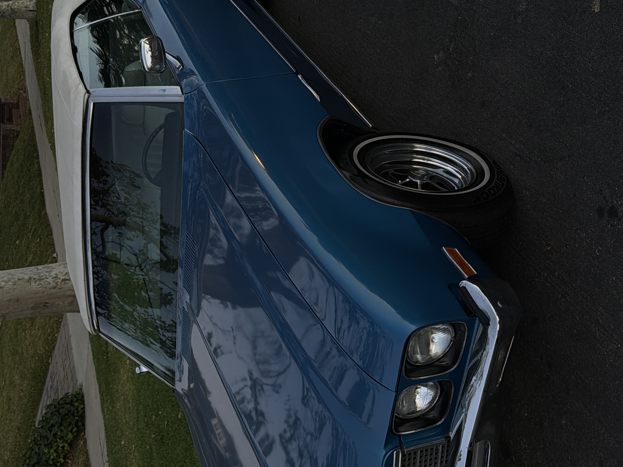 A partially cropped, close-up, medium-shot captures a blue classic convertible car parked on asphalt adjacent to grass and a sidewalk. The car's front end, including the stacked headlights, front bumper, part of the hood, and front passenger-side tire, are prominently visible. The white convertible top is partially visible at the top left corner of the frame. Reflections are visible on the car's polished surface, and a glimpse of the car's interior can be seen through the window. The environment appears to be a residential or suburban setting, likely in Los Angeles, United States of America, based on the style of housing. No people are visible in the frame.
