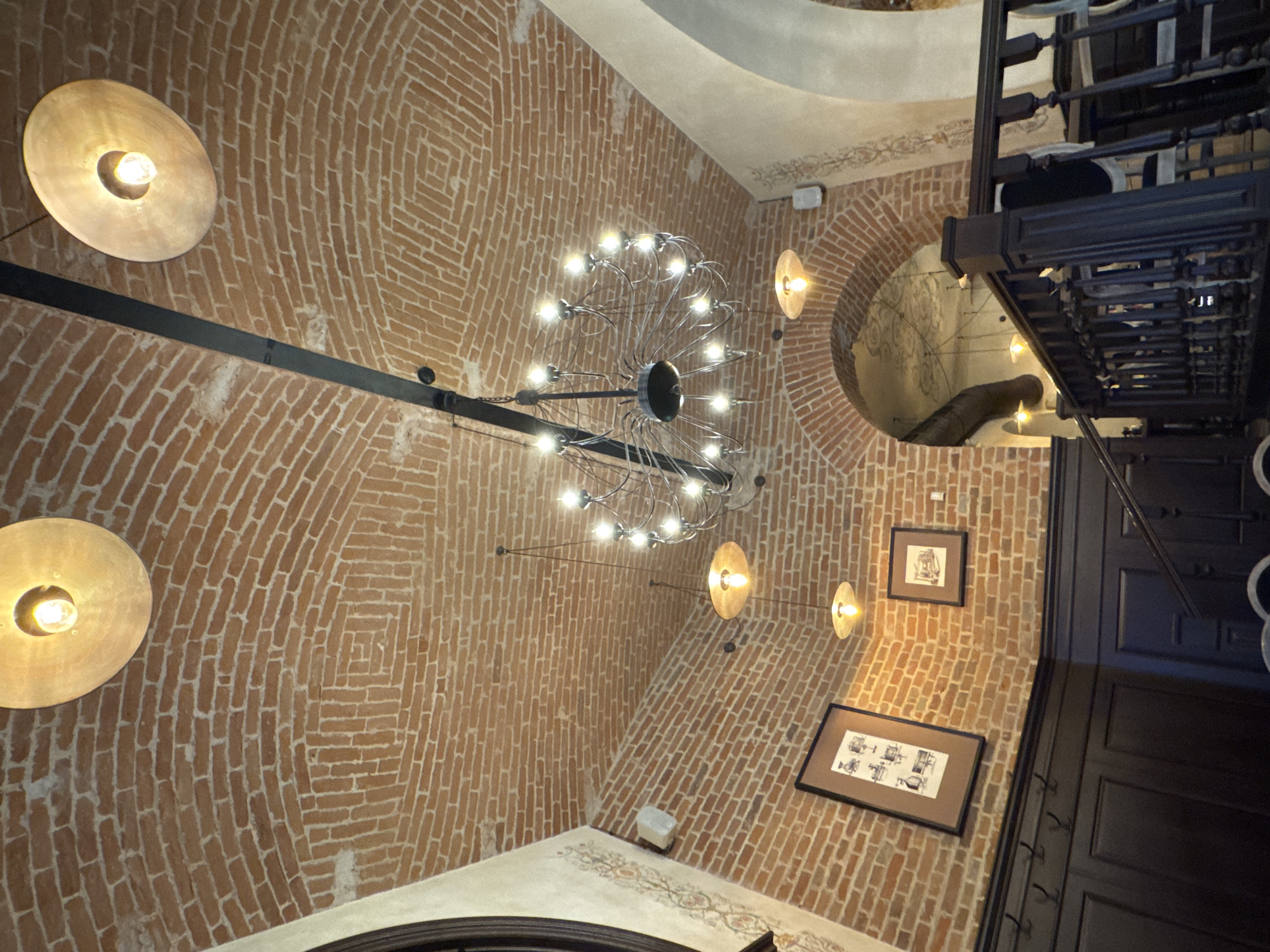This is a low-angle, interior photograph of a room with a brick barrel-vaulted ceiling. The ceiling is decorated with numerous exposed lightbulbs, some in large brass fixtures and others in smaller, pendant lights. A large, ornate chandelier hangs from the center of the ceiling. There are two framed pictures on the brick wall, one larger than the other. A dark wooden railing and stairs are visible in the foreground. The room appears to be dimly lit, with the lightbulbs providing most of the illumination. The architectural style suggests an older building, possibly a historic venue or restaurant. This image was taken in Chernivtsi, Ukraine.