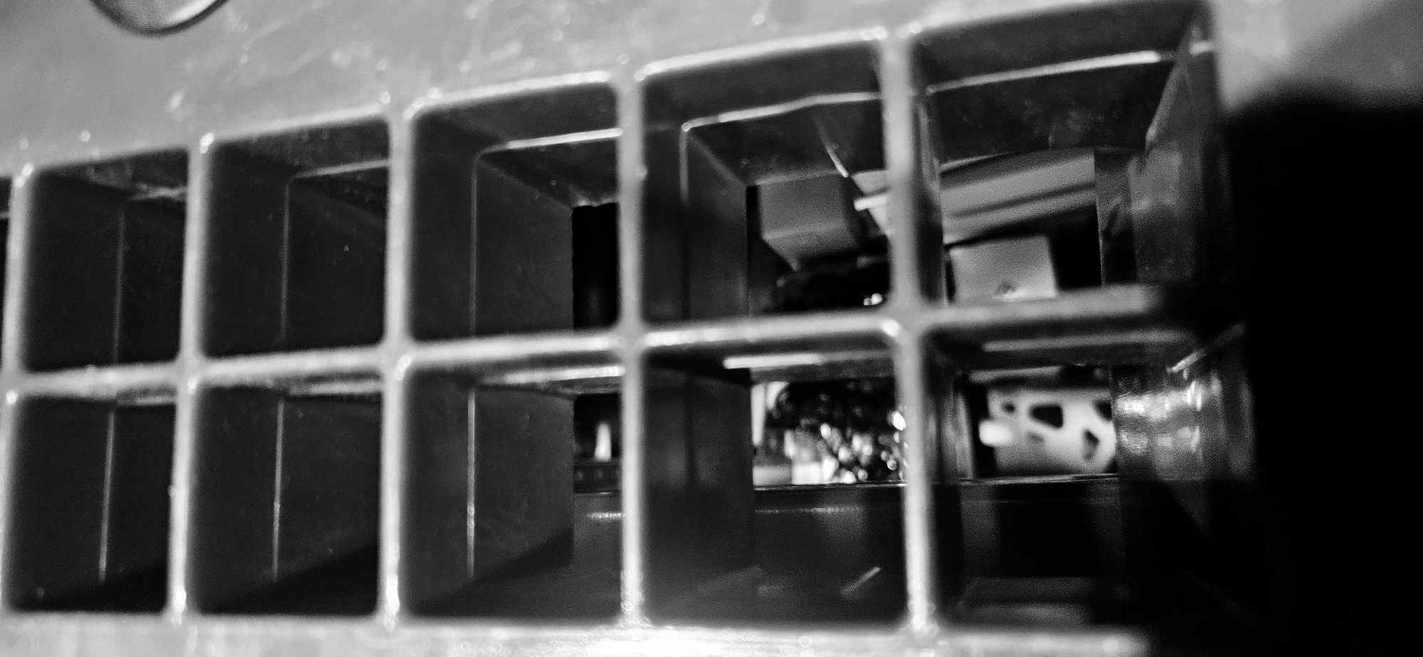 The image shows a close-up view through a grid of rectangular openings. The grid appears to be part of a structure, possibly a vent or grille. Beyond the grid, indistinct shapes and forms suggest the presence of machinery or other objects in the background. The lighting is uneven, with areas of strong contrast and deep shadows. The location is unknown.
