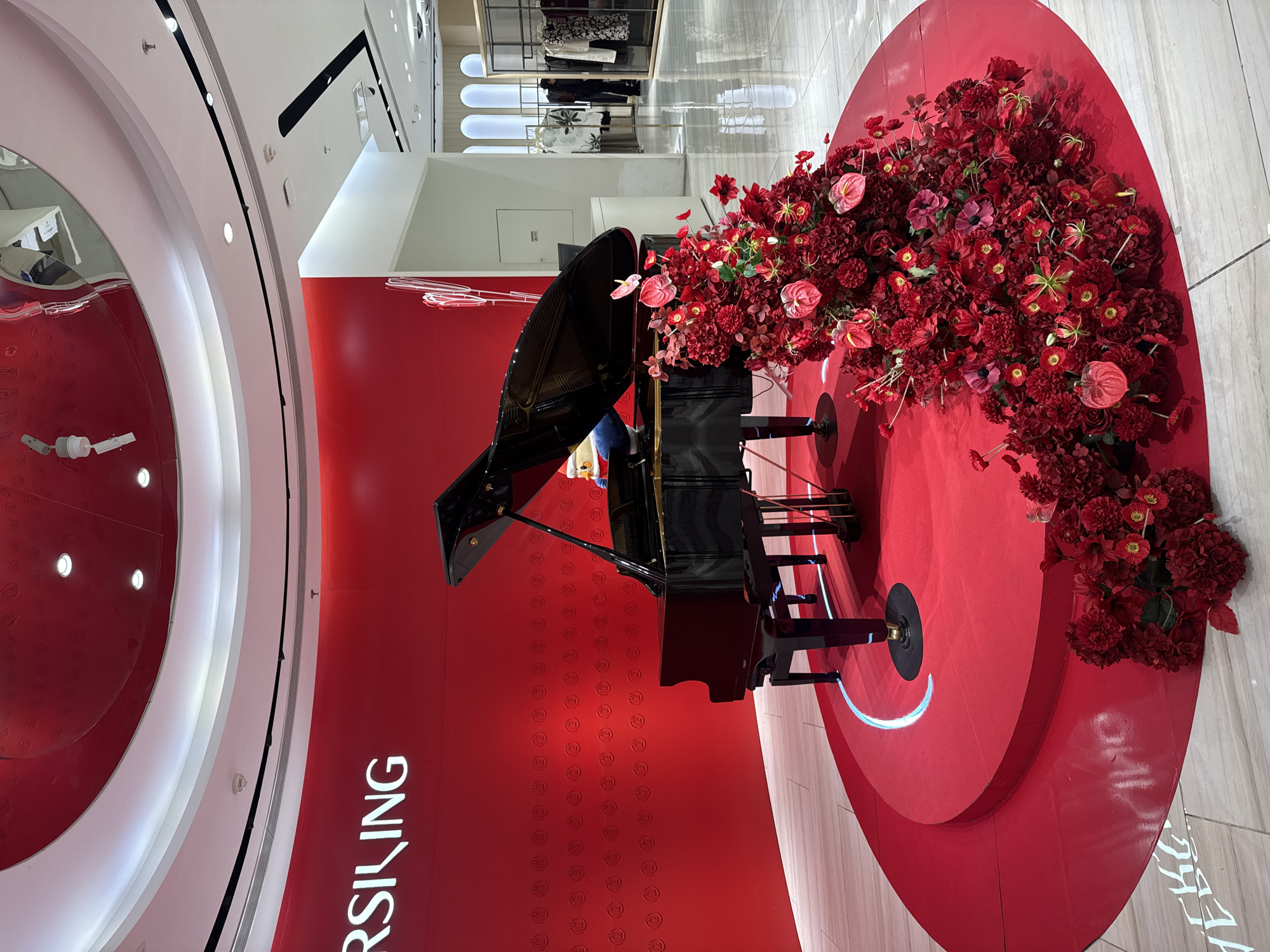 A dimly lit retail interior in Shanghai, China features a black grand piano on a raised red circular platform. The piano is adorned with a dense arrangement of red flowers, and a cartoon character wearing a Santa hat is perched on the keyboard. The background displays a large red wall with embossed circular patterns and illuminated white text that reads "RSILING". To the right of the piano, racks of clothing are visible in a well-lit store section. The floor is a light-colored, polished wood with tiled sections.