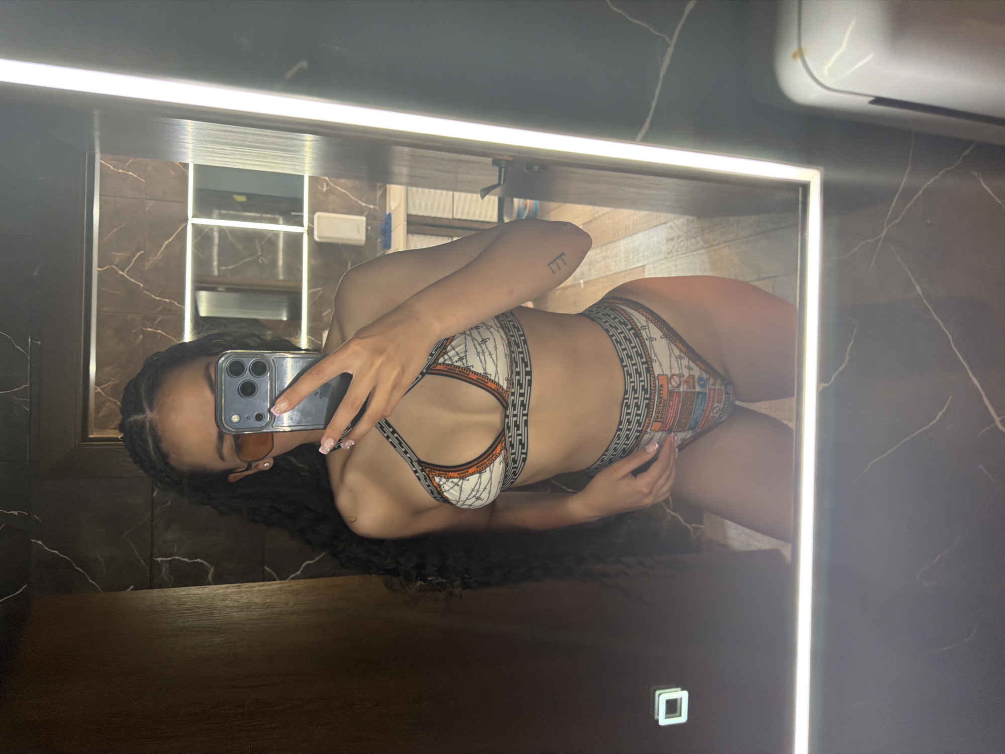 A woman is taking a mirror selfie in a bathroom. She is wearing sunglasses and a bikini. The bikini has a white base with an orange and black geometric pattern and images of text and graphics. The bathroom has dark marble walls and a large, well-lit mirror. The floor appears to be made of wood-look tiles. The overall lighting is bright and modern.