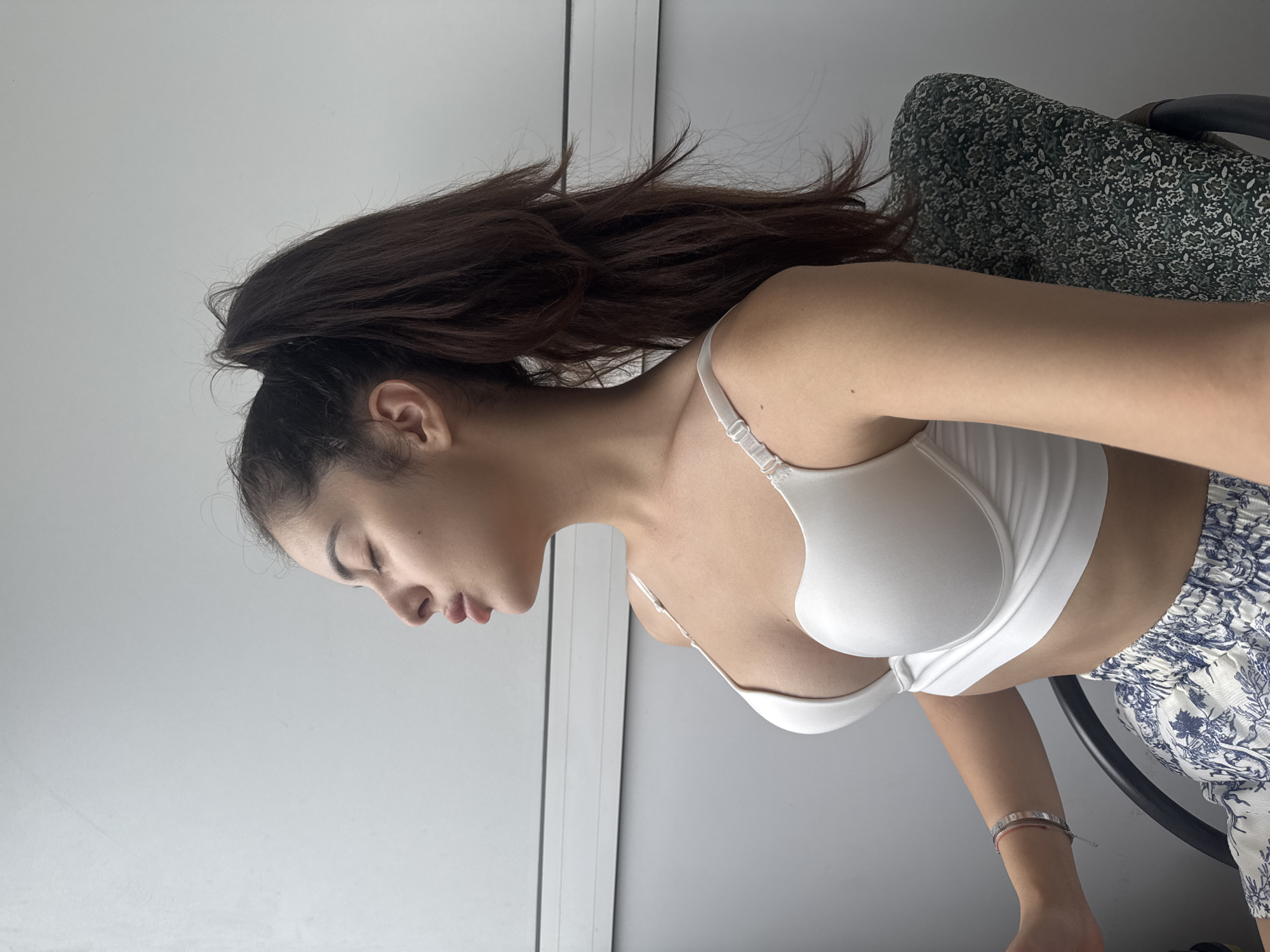 The original image, which is displayed sideways (rotated 90 degrees clockwise), captures a close-up, side profile view of a young woman seated indoors. She has long, dark brown hair pulled back into a high ponytail, with some strands appearing slightly disheveled, suggesting a relaxed style. Her eyes are closed, and her head is tilted gently downwards, conveying a serene or pensive mood.
She is wearing a simple white sports bra or bralette with thin straps, revealing her midriff. Her lower body is partially visible, dressed in light-colored shorts adorned with a distinctive blue and white floral or botanical pattern. On her left wrist, she wears a delicate, thin bracelet, possibly a stack of two, with one appearing to be a metallic band and another a thread-like design.
The background is predominantly a plain, light gray surface, likely a wall or a window, with a thin vertical white line on the left, possibly a door or window frame. Behind her, a portion of a chair is visible, featuring a dark green and black patterned fabric with small floral or leafy motifs. The lighting is bright and even, indicative of daytime indoors, possibly near a window. There are no identifiable location cues within the image itself to suggest Plovdiv, Bulgaria, nor is any text visible. The overall impression is one of calm and quiet introspection.