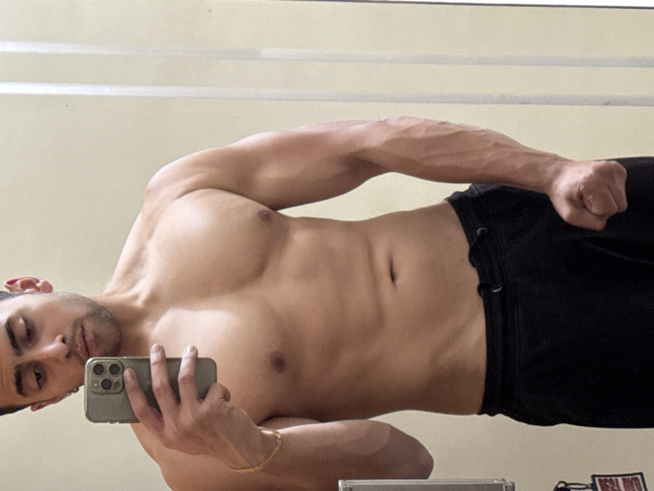 A shirtless man poses for a selfie in a mirror. He's wearing black shorts. The man displays muscular arms and a defined chest. He is holding a smartphone in his left hand. Visible on his left wrist is a gold bracelet. His right fist is clenched. The background is a plain, light-colored wall, and a horizontal line near the top suggests a decorative molding or panel.