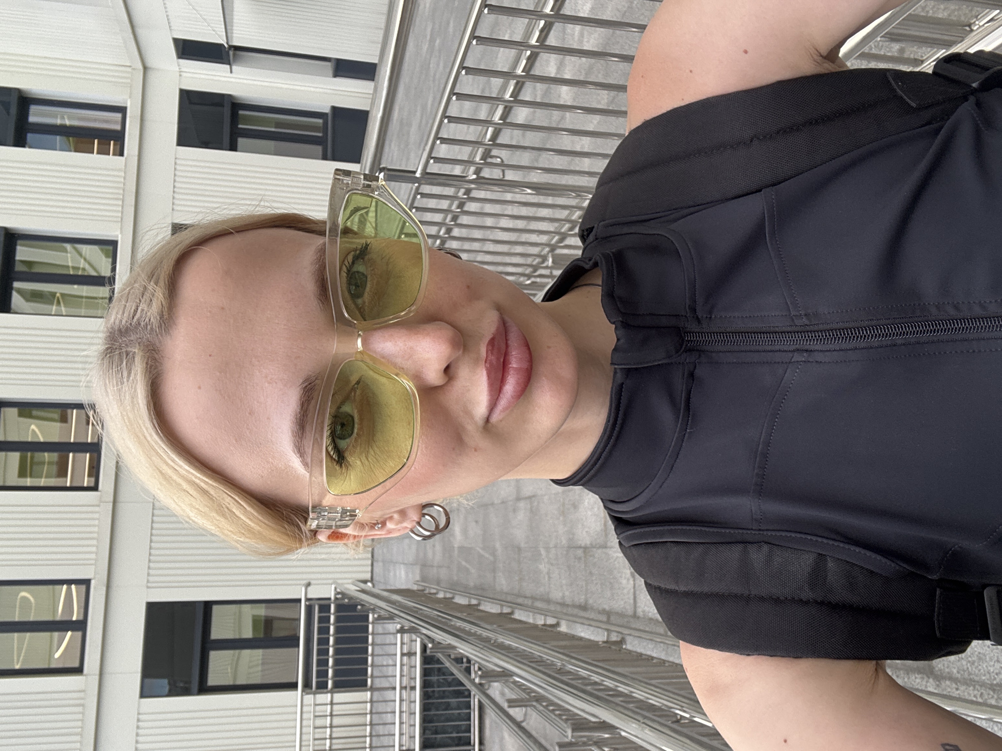 The image shows a close-up, portrait-style shot of a young woman in an urban setting. She has blonde hair and is wearing large, translucent yellow-tinted sunglasses. Her makeup includes visible eyeliner and lip gloss. She is also wearing a black, high-necked vest or jacket with a zipper down the front, and a backpack strap is visible. Large, silver hoop earrings are worn in her left ear.
The background features modern architecture with a facade of horizontal white siding and dark-framed windows. A metal railing, likely part of a staircase or balcony, runs diagonally across the right side of the frame, suggesting an outdoor environment. The lighting indicates it is daytime, possibly overcast, as there are no strong shadows. The overall impression is a casual, confident pose. The location context is Moscow, Russia. No text is visible in the image.
