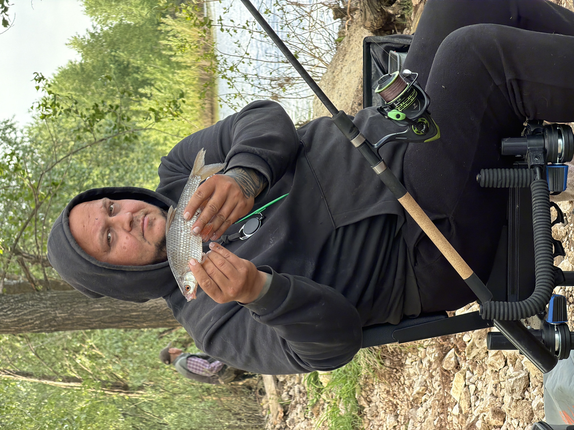 A man in a black hoodie is sitting in a fishing chair and holding a fish. He is outdoors near a body of water and trees. A fishing rod is set up next to him. Another person is visible in the background. The ground is covered in rocks and dirt. The location is Noviy, Russia.