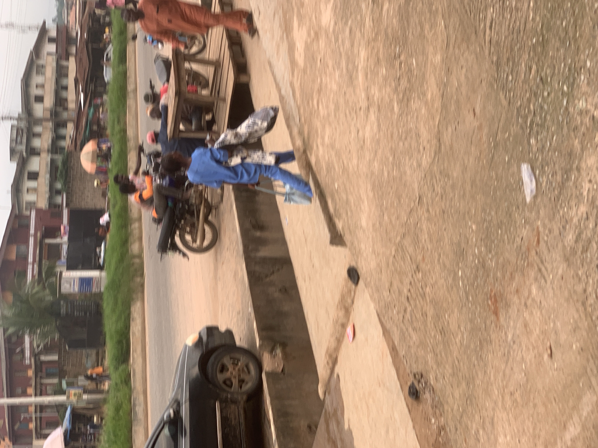 In Shagamu, Nigeria, a street scene unfolds with a young person in a blue outfit walking with a light blue bag and a bundle of patterned fabric. They are moving from right to left across a sandy, textured surface. To the left, a black car is parked. In the background, a motorcycle is visible, with a person in an orange outfit standing near it. Further in the background, several buildings, some with red roofs, line the street, and a few scattered trees and patches of green vegetation are present. The overall environment appears to be a busy, possibly commercial area.