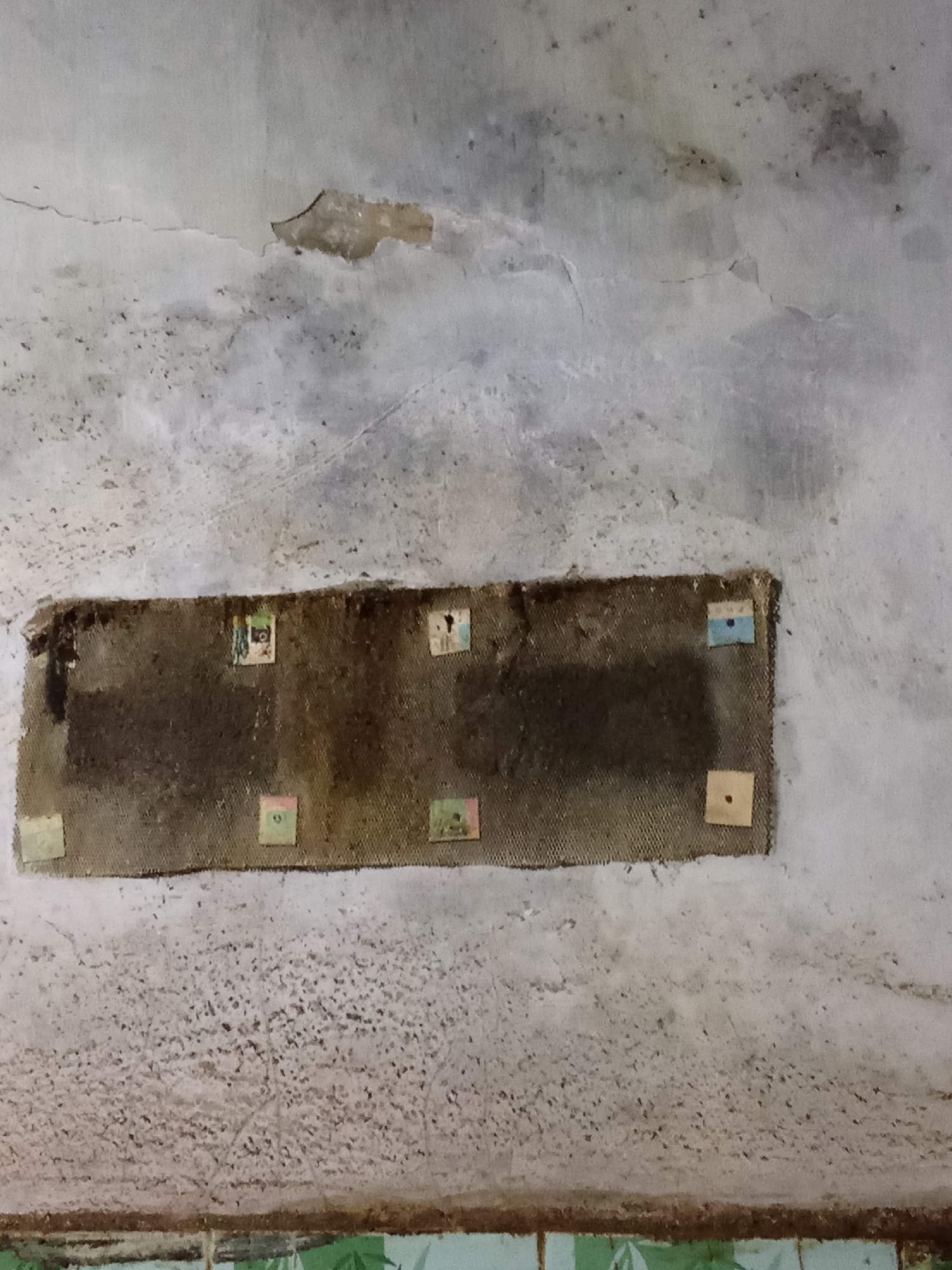 The image depicts a section of a rough, weathered concrete wall in what appears to be an indoor or sheltered outdoor setting. The wall is cracked and stained, with patches of peeling plaster and dark mold or water damage. A rectangular area of coarse, dark brown fabric, possibly burlap or hessian, is affixed to the wall. This fabric covers a darker, possibly recessed, area or opening. Scattered across the fabric are several small, square-shaped pieces of paper or cardstock, each featuring a colorful image or pattern. These appear to be adhered to the fabric. Below the fabric, the wall exhibits a textured, granular surface. The very bottom of the image shows a sliver of a tiled surface with a green and white patterned design. The lighting is dim and appears to be artificial. There are no people or animals visible in the image. The context of Pemalang, Indonesia, suggests a potentially humid climate, which could explain the presence of mold and the weathered appearance of the wall. However, without further information or context, it is impossible to definitively pinpoint the exact location within Pemalang.