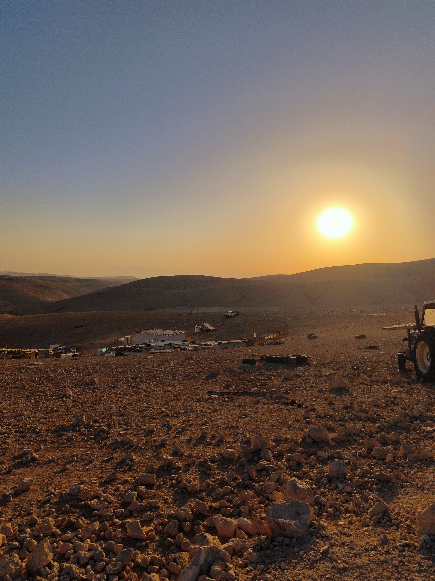 This is a vertical, eye-level, medium shot taken during sunset in the Palestinian Territories. The sun is low on the horizon, casting a warm, orange glow over the rocky, arid landscape. The foreground is dominated by numerous stones and pebbles scattered across the reddish-brown earth. In the mid-ground, a cluster of low-lying, simple structures, possibly temporary dwellings or storage sheds, are visible. A white vehicle is parked to the left of these structures, and various items, including tires, are strewn around the area. A tractor is partially visible on the right side of the image, with its rear wheel prominent. Rolling hills form the background, receding into the hazy distance under a gradient sky that transitions from a pale blue at the top to a bright orange near the horizon. The overall impression is one of a remote, undeveloped settlement in a desert environment at the end of the day. The location is identified as the city of Keidar, Palestinian Territories. There are no visible people in the image.