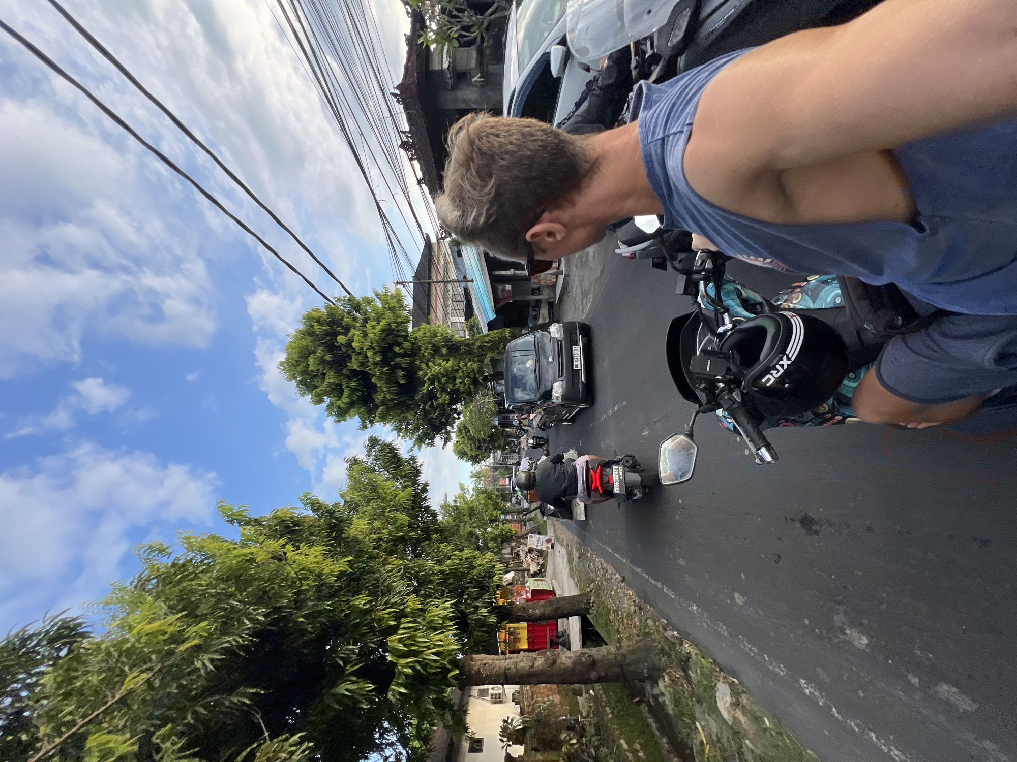 The image depicts a man, likely the photographer, on a motorcycle in Sayan, Indonesia. The scene is an urban street with traffic consisting of motorcycles and a black car. Vegetation lines the street on both sides, including mature trees. Above, power lines crisscross the sky with scattered clouds. The man is wearing sunglasses and a blue shirt. A helmet with the branding "XRC" is visible on the motorcycle. The photograph is taken at an angle, giving a distorted perspective of the environment.
