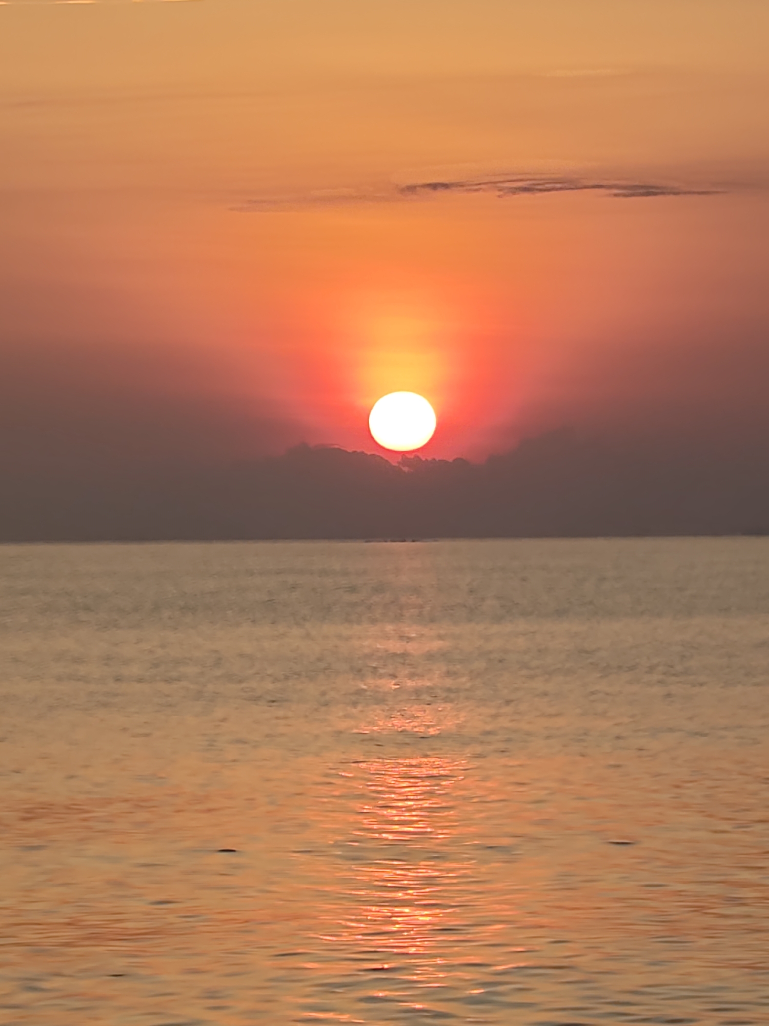 The image displays a coastal scene in Lhokseumawe, Indonesia, captured during either sunrise or sunset. The sky, occupying the upper half of the vertical frame, exhibits a gradient of warm colors, transitioning from orange at the top to vibrant red closer to the horizon. A prominent, intensely bright white circular sun is centered horizontally, positioned just above the horizon line, partially obscured by a band of dark, horizontally elongated cloud formations directly beneath it.
The sun's light permeates the lower sky and casts a distinct, shimmering, reddish-orange reflection path across the calm surface of the water, which comprises the lower half of the image. The water surface shows minor ripples, reflecting the ambient light. A faint, dark, horizontal line is discernible on the distant horizon, suggesting a remote landmass or large structure. No human subjects, animals, or identifiable man-made objects are present in the foreground or midground. The scene is static, with light interaction between the celestial body, atmosphere, and water.