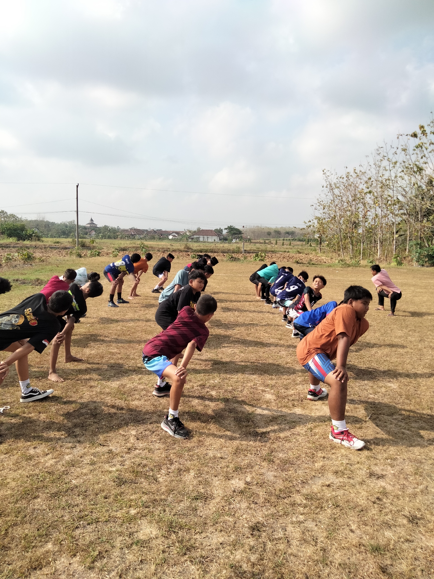 The image depicts a group of young males engaged in a physical training exercise outdoors on a dry, grassy field. They are arranged in a line, facing away from the camera, and are performing a bend-over stretch with their hands on their knees. The background reveals a rural landscape with sparse trees, a few scattered buildings in the distance, and utility poles. The sky is overcast with clouds. The scene appears to be set in Pucakwangi, Indonesia, based on the context. The participants are dressed in casual athletic wear, including t-shirts, shorts, and athletic shoes. The overall atmosphere suggests an organized group activity, possibly a sports practice or physical education class.