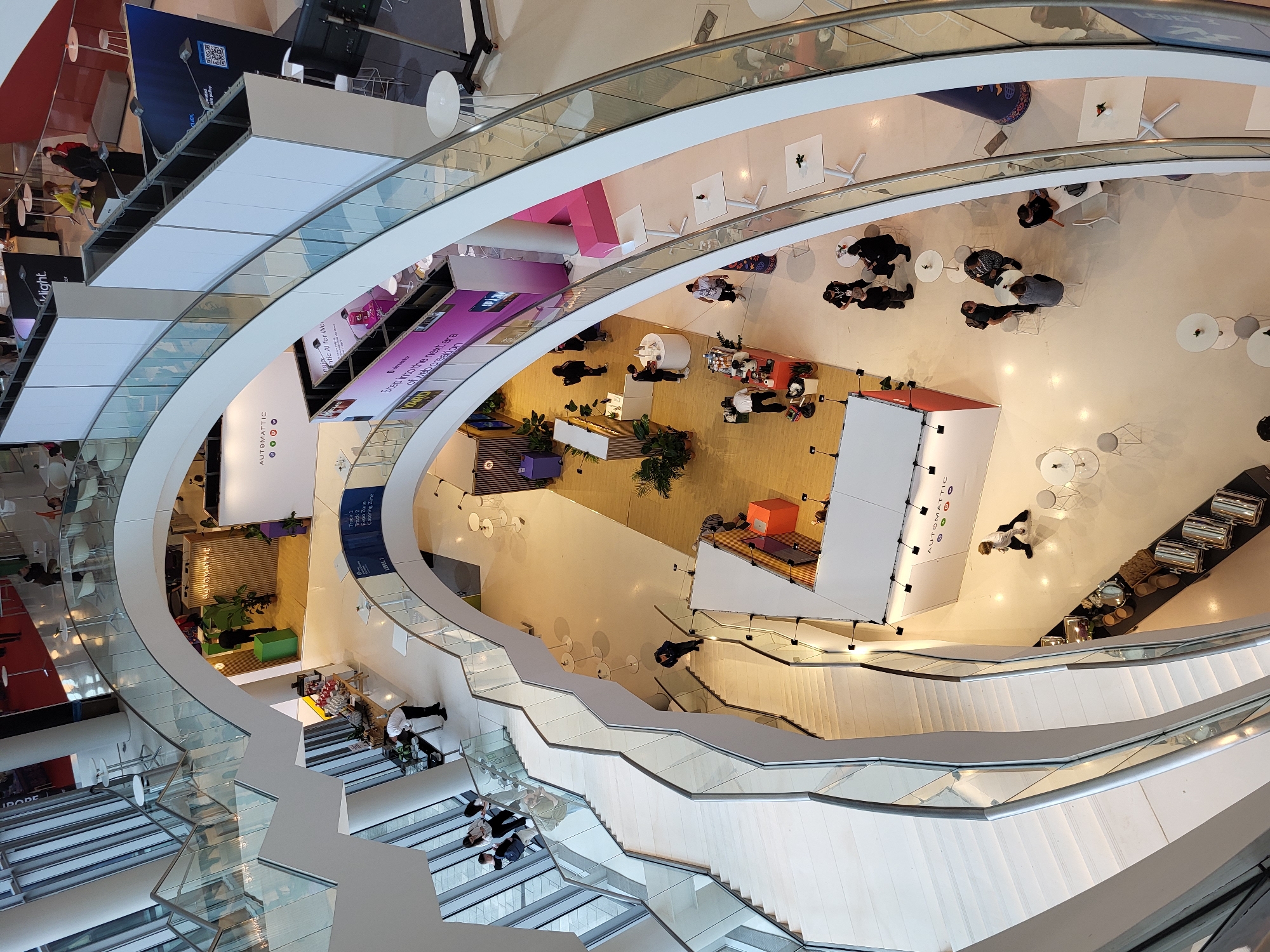 This high-angle, downward-facing photograph depicts a multi-level, modern indoor event venue, possibly an exhibition hall or conference center atrium. A prominent white, winding staircase with glass railings curves through the central space, connecting at least three visible levels.
On the ground level, a light-colored floor hosts several activity zones. A large "AUTOMATTIC" branded booth, primarily white with a red base, is in the foreground right. Another "AUTOMATTIC" display with green accents and potted plants is further back on the left. A purple and pink booth promotes "Intelic AI for Work" and "Step into the next era of web creation." A central section of the floor features light wood paneling, where a barista operates a coffee station and a person in a white top attends to a circular display counter. Catering stations with silver chafing dishes are arranged on a black counter to the far left. Numerous white and light gray round and square tables with matching chairs are distributed across the open areas, occupied by individuals and small groups engaging in conversation or resting. A blue sign indicates "LEVEL 1" and lists "Track 1," "Track 2," "Expo Zone," and "Catering Zone."
Intermediate levels consist of curved walkways with glass railings, also furnished with white tables and chairs. On the uppermost visible level, a red carpeted area contains additional tables and chairs, with white display walls bearing partial branding such as "darklight." and "CLICK," the latter displaying a QR code.
Approximately 25 individuals are observed throughout the scene, engaged in activities including walking, standing in groups, sitting at tables, and working at service stations. The environment is brightly lit with white walls, polished floors, and large vertical windows on the left allowing natural light into the structure. The overall aesthetic is clean, modern, and open.