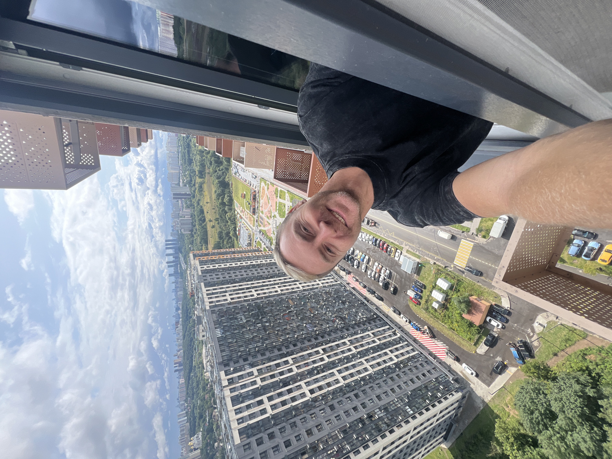 In the city of Khimki, Russia, a man with blond hair and a beard, wearing a black t-shirt, takes a selfie from a high vantage point, possibly a balcony or window. He is smiling and looking at the camera. Below him is a large, modern residential building with a grey facade and numerous windows. The surrounding area features a parking lot with several cars, green spaces, and other buildings in the distance, indicative of an urban environment. The sky is partly cloudy.