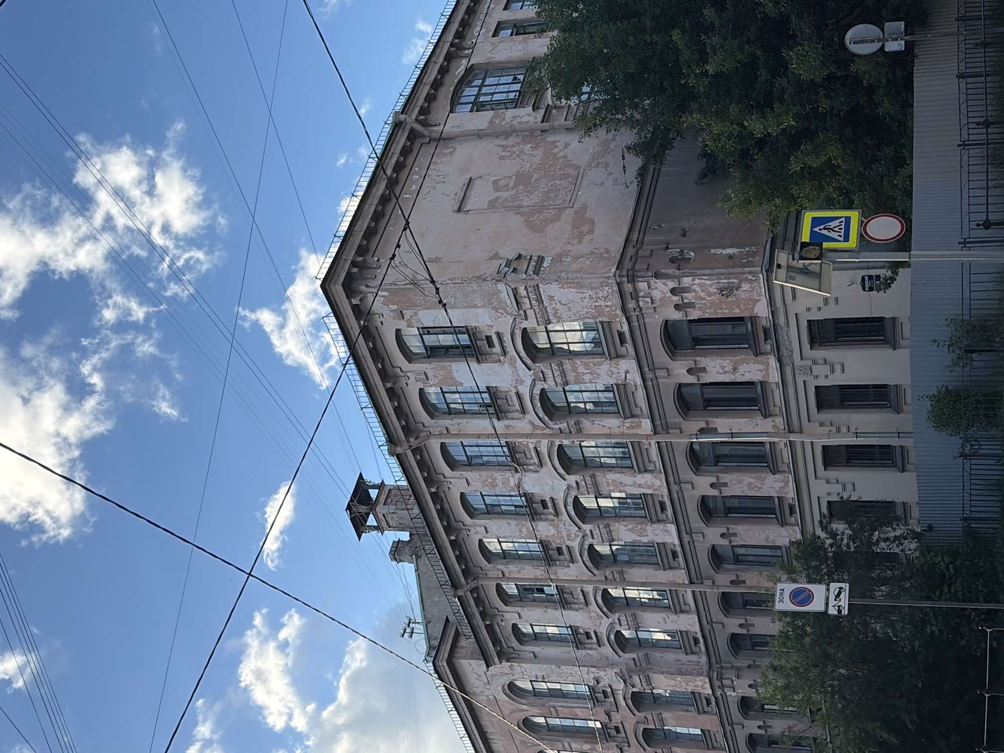 The image captures a street scene in Saint Petersburg, Russia, featuring a large, aged building with a pale pink facade exhibiting peeling paint. The architecture suggests a historical industrial or residential structure, characterized by multiple rows of arched windows, many of which appear to be old or damaged. A prominent ventilation shaft or small tower is visible on the roof. Overhead, a network of power or trolley wires crisscrosses the sky, which is partly cloudy with a bright blue sky peeking through.
In the foreground, a street sign pole displays several signs. The topmost sign is a circular blue sign with a red border and a diagonal red line, indicating "no parking" or a restricted zone, with Russian text above it reading "ЗОНА" (ZONA - ZONE). Below this is a rectangular sign depicting a tow truck towing a car, further emphasizing parking restrictions. Further to the right, another pole holds a yellow square sign with a blue border and a white pedestrian symbol, indicating a crosswalk. A red and white circular sign is mounted below it.
The building is bordered by a grey metal fence in the middle ground. Lush green trees are visible to the right of the building and behind the fence, suggesting a presence of greenery within the urban environment. No people are visible in the frame. The overall impression is one of an urban setting with a blend of old architecture and functional street infrastructure.