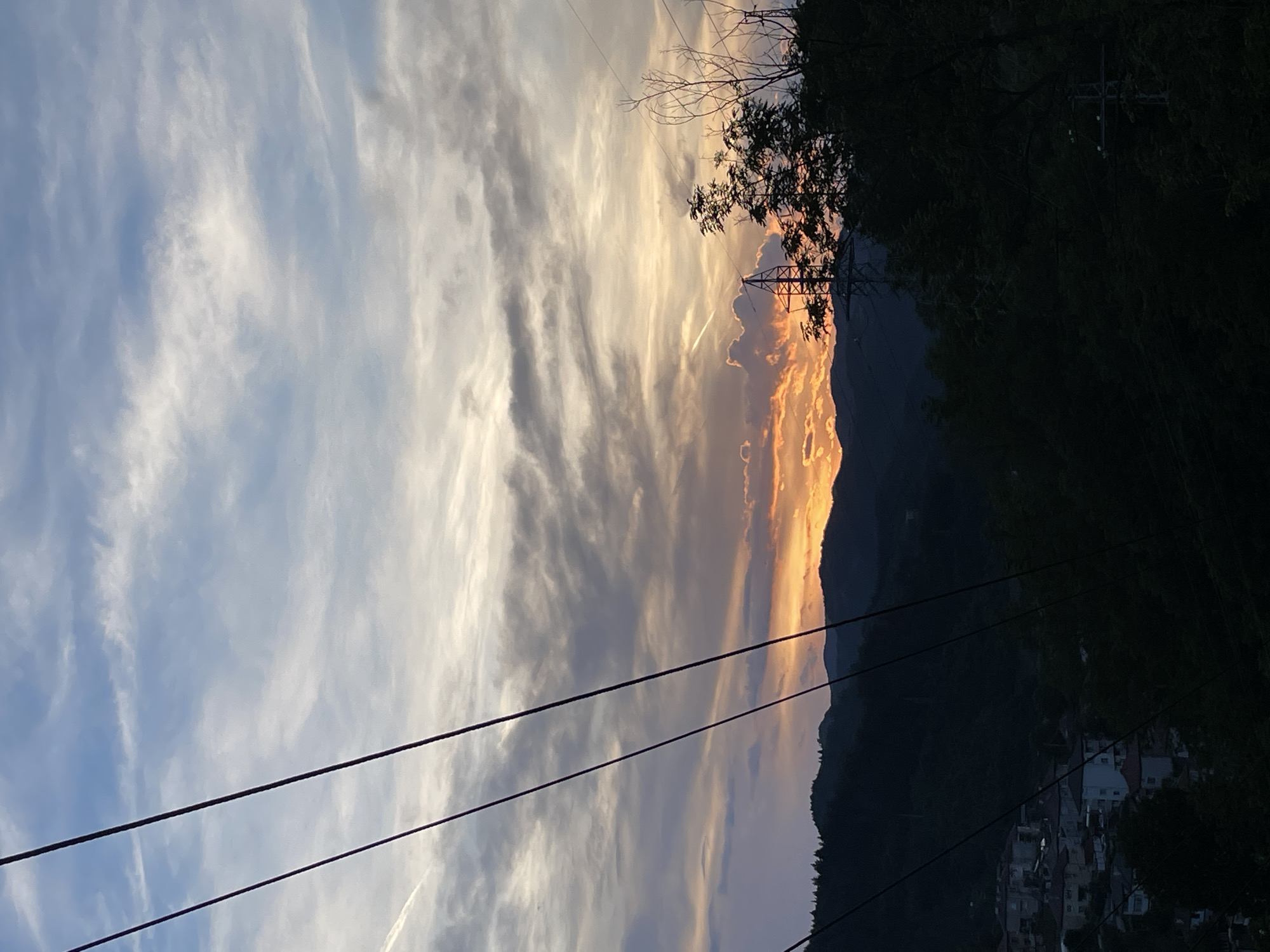 The image depicts a sunset over Zlatograd, Bulgaria. The sky is filled with dramatic clouds, tinged with orange and yellow from the setting sun, contrasting with cooler blues and grays. In the foreground, silhouetted against the vibrant sky, are several power lines stretching across the frame, casting dark shadows. Beyond the power lines, rolling hills covered in dark green trees are visible. Nestled in the valley between the hills, a cluster of houses with red-tiled roofs forms a small village. The scene is a picturesque blend of natural beauty and human habitation, captured during the twilight hours.