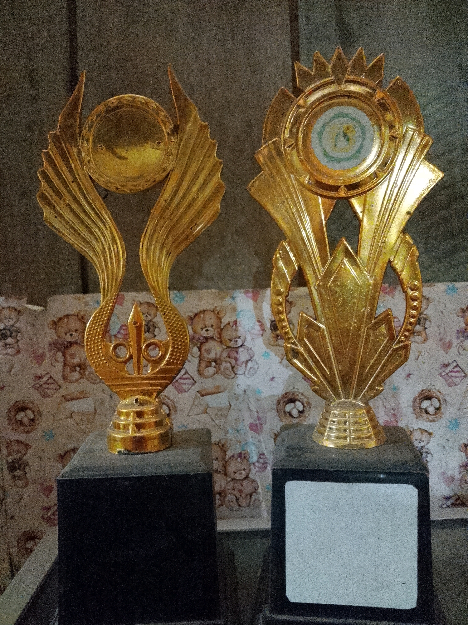 Two gold-colored trophies are displayed on dark grey rectangular bases. The trophy on the left features a circular medallion at the top, flanked by ornate wing-like structures. The trophy on the right has a sunburst-like crown above a circular emblem containing concentric rings of blue, green, and yellow. Both trophies are made of a metallic material and have a glossy finish. The background consists of a patterned fabric with images of teddy bears and other motifs, and a textured grey wall. The scene appears to be an indoor setting, possibly a room in Wadang, Indonesia. There are no people visible in the image.