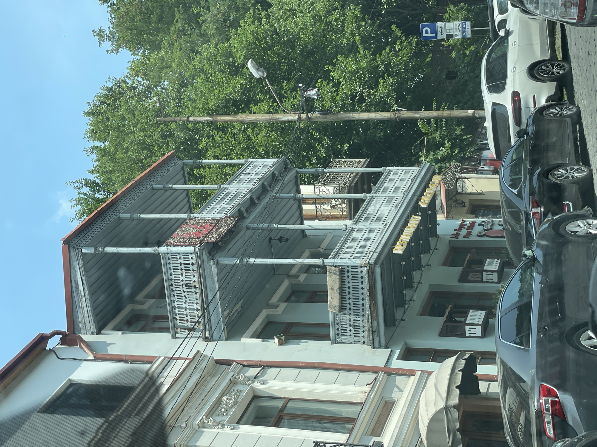 A light blue building with three balconies is featured in this image, located in Tbilisi, Georgia. The balconies are ornately decorated with wooden balustrades and are adorned with rugs. The building also has a red-tiled roof. Next to the building, a dark gray car is parked, and further down the street, a white SUV is visible. A utility pole with wires and a lamppost stands between the building and the street. Lush green trees form a dense backdrop behind the building and the utility pole. The sky above is a clear, bright blue. A parking sign with a blue "P" symbol is also visible.
