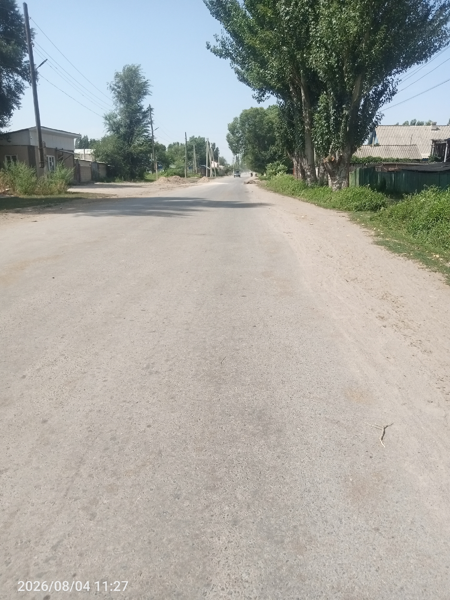 The image depicts a street in the city of Djany-Pahta, Kyrgyzstan, on August 4, 2026, at 11:27 AM. The street is paved with asphalt and appears to be a two-lane road with no visible lane markings. The left side of the road is bordered by a single-story building with a light-colored facade and a fence, surrounded by vegetation. Further down the street, more buildings and trees are visible. The right side of the road features a line of mature trees with dense green foliage, casting shadows on the asphalt. Beyond the trees, a residential property with a grey-tiled roof and a green fence is visible. The environment suggests a residential or semi-rural area within the city. The sky is clear and blue. A few vehicles are faintly visible in the distance. The overall scene is one of a typical day in this location.