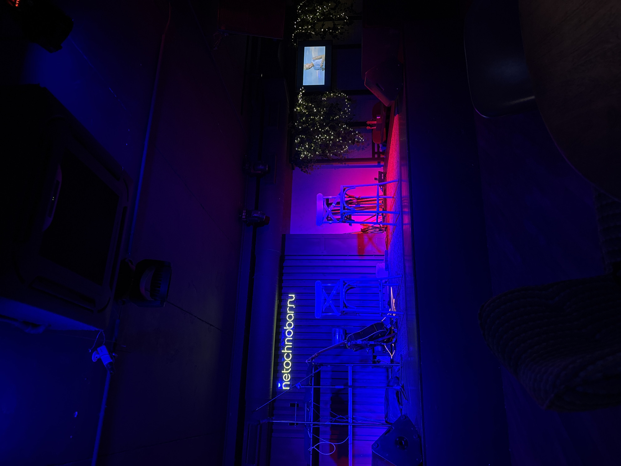 The image shows the interior of a bar in Krasnoyarsk, Russia. The bar's name, "netochnobarru," is displayed vertically in neon blue on a wall. A stage area is lit with blue and pink neon lighting. There is musical equipment set up on the stage. There is a TV screen with an image displayed on the wall. String lights are hung around plants. The overall lighting is dim and atmospheric.