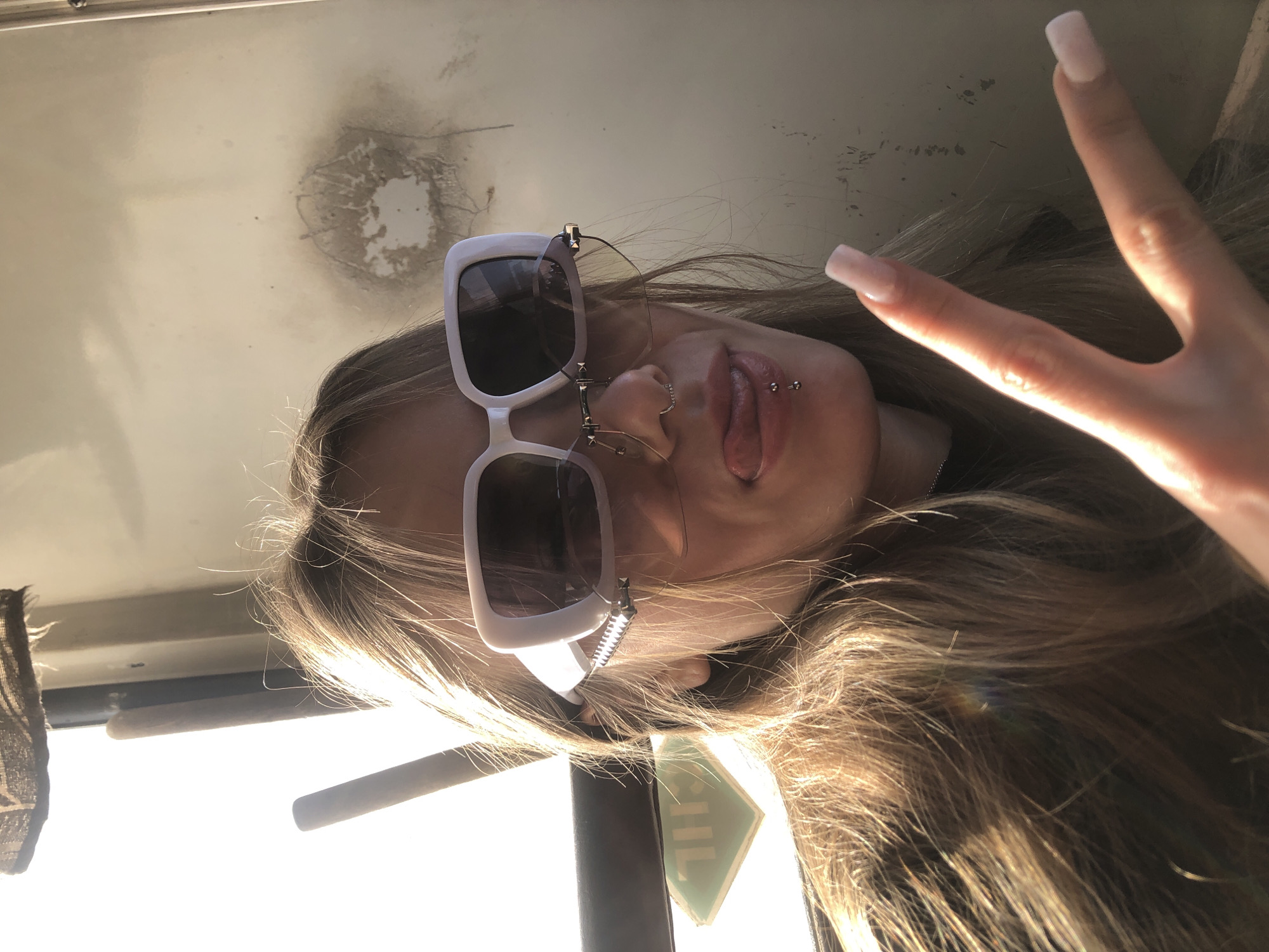 This close-up portrait captures a young individual, identified as being in the city of Foki, Russia, within what appears to be the interior of a vehicle or building, likely near a window. The subject, a female with long, light brown hair illuminated by strong backlight, is centered in the frame. She wears large, white, rectangular sunglasses, beneath which a second pair of rimless glasses is visible. Her facial expression includes an extended tongue, curled slightly upward, and she has a silver hoop piercing in her septum and a barbell piercing in her lower lip. Her right hand is raised, displaying a "peace" sign with two fingers extended; her fingernails are long, square, and light pink.
The background on the left features a bright window emitting strong sunlight, a dark, textured fabric hanging from above, and a vertical cylindrical handle or pole. A green, diamond-shaped sign with inverted light lettering is visible through the window. The background on the right consists of a plain, light-colored panel or wall, exhibiting a prominent dark, circular smudge with a lighter center, and several smaller dark marks.