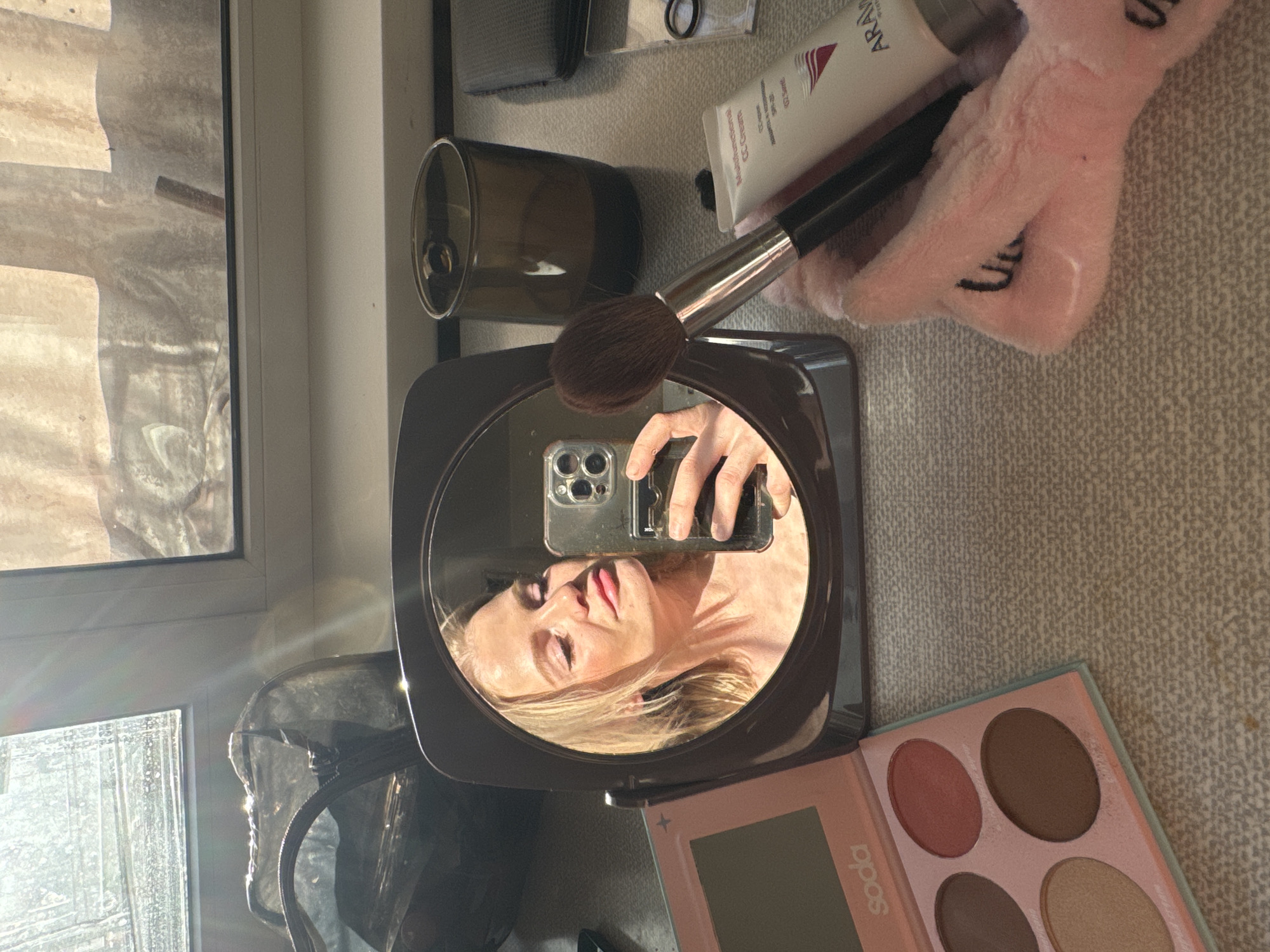 A woman's reflection appears in a small makeup mirror, held by her left hand. The camera is a modern smartphone. The surrounding objects suggest a personal grooming or makeup application context. These objects include a makeup brush, a tube of lotion (labeled "ARAVIA Professional"), a plush pink object with lettering, a transparent cosmetic bag, a dark-colored drinking glass, a small storage container, and a makeup palette. The palette is labeled "SODA." The presence of window frames and intense light suggests the setting is indoors, near a window. The location within Saint Petersburg, Russia is not deducible from these details.