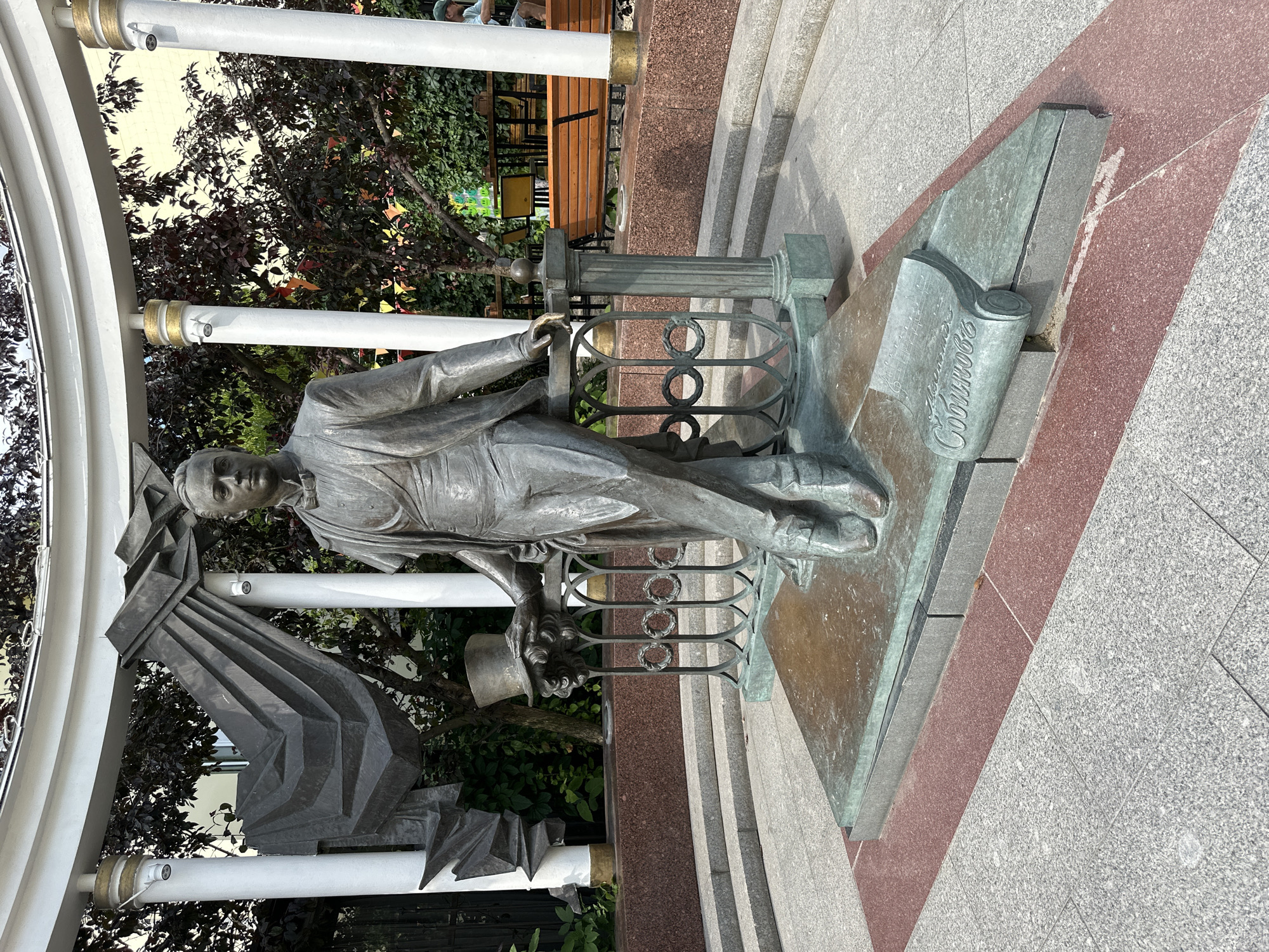 The image shows a bronze statue of a man in formal attire, holding a top hat in his left hand and resting his right arm on a decorative railing. The statue is positioned on a triangular bronze base that is set on a multi-tiered pedestal. To the left of the statue, a large abstract metal sculpture is integrated with the surrounding white, cylindrical columns and a curved white canopy. On the right side of the base, a rolled-up scroll with musical notation is depicted. The surrounding area is paved with granite tiles and red granite borders. In the background, there are trees with dark foliage, a building with yellow walls, and outdoor seating with benches. A person in a green cap is visible in the far right background. The scene appears to be an outdoor public space, possibly a park or square, in the city of Putyatino (Semivragovskaya S/A), Russia.