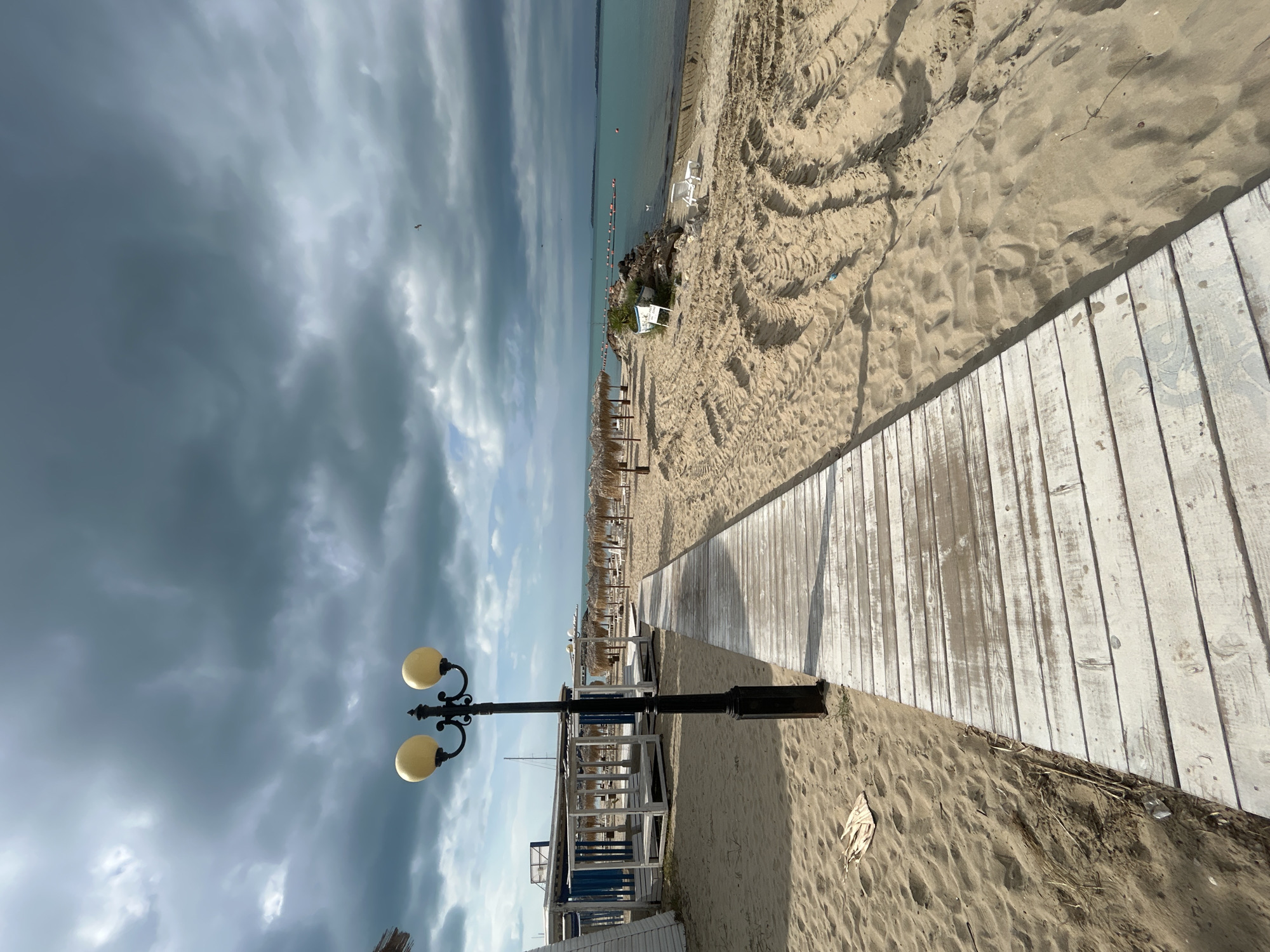 The image depicts a beach scene in Sveti Vlas, Bulgaria. A weathered white wooden boardwalk extends from the bottom right towards the center of the frame, leading towards beach umbrellas and lounge chairs arranged along the shore. The beach is sandy with tire tracks visible. A vintage-style lamp post with two globe-shaped lights stands at the edge of the boardwalk. The sky is overcast with dark, heavy clouds. The sea is calm and a light blue-green color. A few people are visible in the water. White structures possibly associated with a beachside establishment are partially visible on the left, partially obscured by the lamp post.