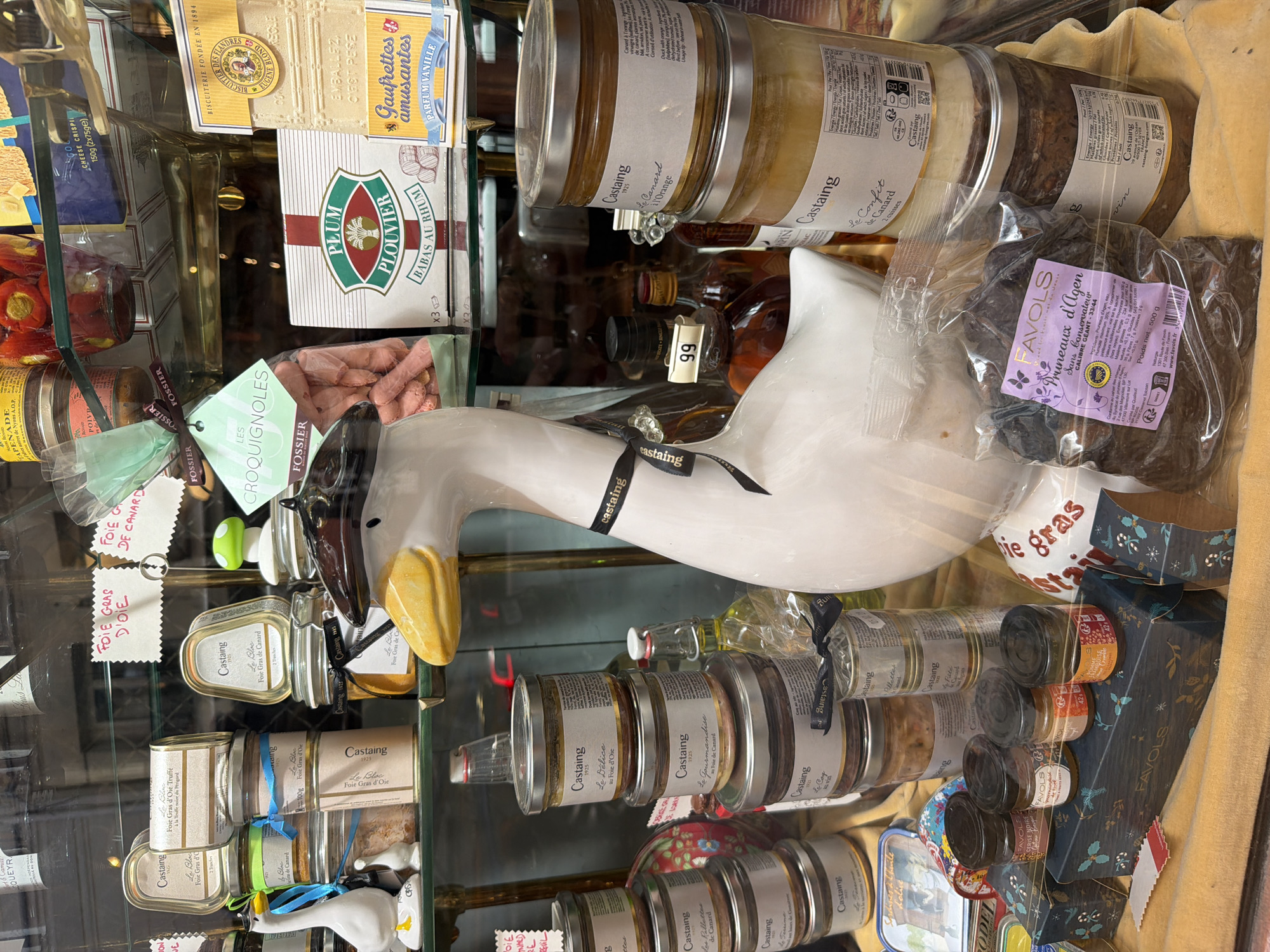 The image shows a shop window display in Paris, France, filled with gourmet food products. There are several jars and cans labeled "Castaing," indicating foie gras and other duck-based products. Additional items include "Favols" pruneaux d'Agen, gaufrettes amusantes, and babas au rhum. A ceramic duck figurine with a black ribbon and hat is positioned centrally in the display. A "99" price tag is visible. The overall impression is of a specialized food shop offering regional French delicacies.