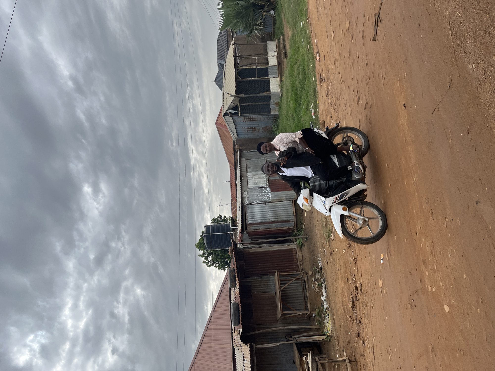 Three young men are posing on a white motorcycle on a dirt road in what appears to be a rural Nigerian setting. The man in the middle is wearing a light pink shirt, and the other two are wearing black. The man on the far right is smiling and giving a peace sign. The motorcycle is white and has a sticker on the side. Behind them are several rustic buildings with corrugated metal roofs and walls. One building has a large black water tank on its roof, suggesting a village or community setting. The ground is reddish-brown dirt, and there is some sparse green grass and scattered debris. The sky is overcast with dramatic, cloudy formations, indicating it might be late afternoon or an impending storm. The overall mood seems relaxed and friendly, with the subjects appearing to enjoy their moment together. There is no visible text in the image.