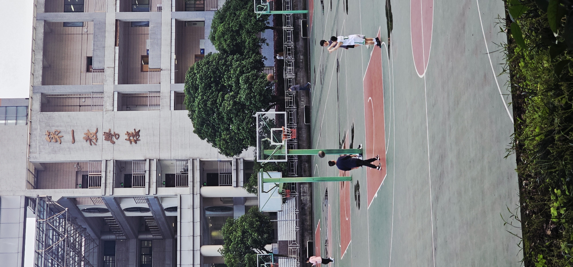 A horizontally oriented, medium-angle, full outdoor shot depicts a basketball court located next to a tall building. The building, situated on the left side of the image, features a facade with Chinese lettering.
Several people are present on the basketball court, some actively playing basketball. One individual is mid-throw with a basketball, while another is standing near a basketball hoop. Two other individuals are standing closer to the top of the scene.
The environment includes trees interspersed throughout the scene, adding a natural element to the urban setting. The basketball court itself is lined with green, white, and red paint. The overall lighting suggests it is daytime. The location of this scene is Yuzhong District, China.