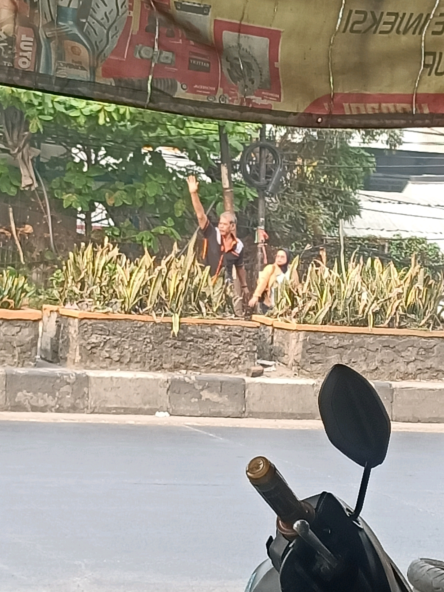 The image captures a street scene in Gandasari, Indonesia, likely taken from a vehicle. In the foreground, the handlebars and side mirror of a scooter are visible. Beyond the scooter, a raised concrete barrier lines the street, and behind it, a planter box filled with tall, green and yellow striped plants is present. Two individuals are standing near the plants. A man in a black and orange shirt with his right arm raised in a wave is on the left. To his right, a woman wearing a yellow jacket and dark pants is also standing. In the background, there are trees and some buildings. A banner with an image of what appears to be a red vehicle and text is stretched across the top of the scene.
