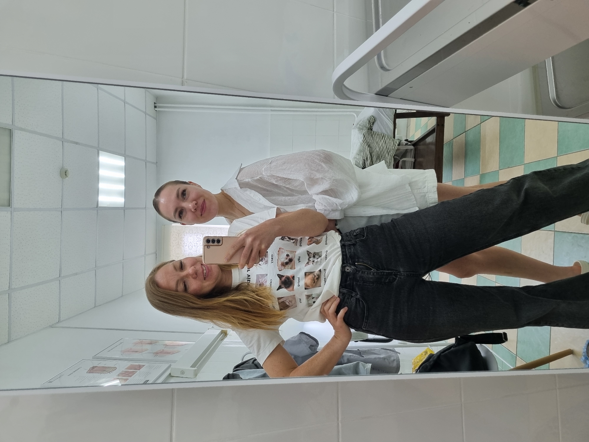 The image shows two young women taking a mirror selfie in what appears to be a medical or examination room. The woman on the left is wearing a white t-shirt with a print of cats and black high-waisted jeans. She is holding a smartphone to take the photo. The woman on the right is wearing a white oversized button-down shirt and is standing behind the first woman. The room is tiled with white and light green squares on the floor and white tiles on the walls. There are medical charts on the wall and a table with a striped blanket in the background. The room is well-lit by fluorescent ceiling lights.