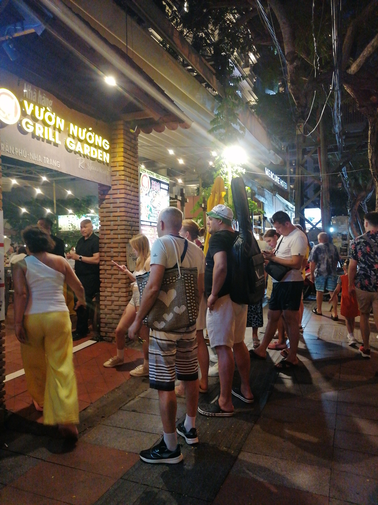 The image captures a nighttime street scene in Nha Trang Ward, Vietnam, in front of the "Vườn Nướng Grill Garden" restaurant. The restaurant's illuminated signage is visible. The exterior has a brick facade. A few trees are visible behind the restaurant. Power lines extend above the restaurant, partly obstructing the view.
A diverse group of people occupies the sidewalk. Several individuals are standing or walking, some using mobile phones. There is a guitar case being carried by a person in a black shirt. Others are wearing shorts and summer clothes. The sidewalk is paved with gray tiles. Shadows stretch across the sidewalk, indicating artificial light sources, including a sign for the Mercure Hotel in the background. The general environment suggests a lively, tourist-oriented atmosphere.