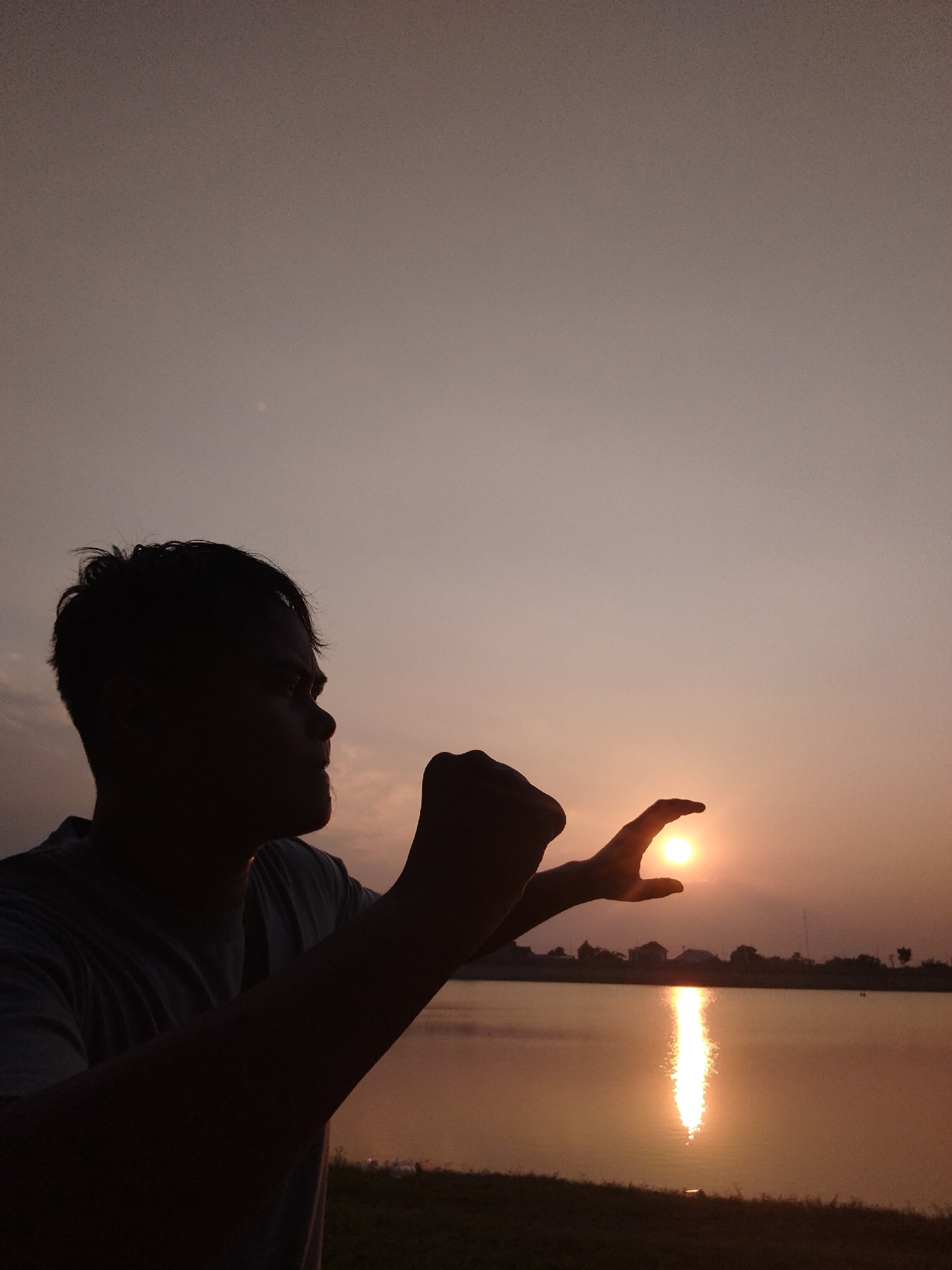 A silhouetted man stands with his profile facing right, his right hand reaching out and cupping the sun. The sun is a bright orb of orange light, positioned between his thumb and forefinger, casting a reflection on the water's surface. The man is wearing a t-shirt. He is positioned in the foreground, with a body of water and a distant treeline visible behind him. The sky is a gradient of pale orange and grey, indicative of a sunset. The overall scene suggests a location near water, possibly a lake or river, with some buildings and trees on the horizon. Given the context of Sambirejo, Indonesia, this could be a natural landscape or a man-made reservoir in or around the area.