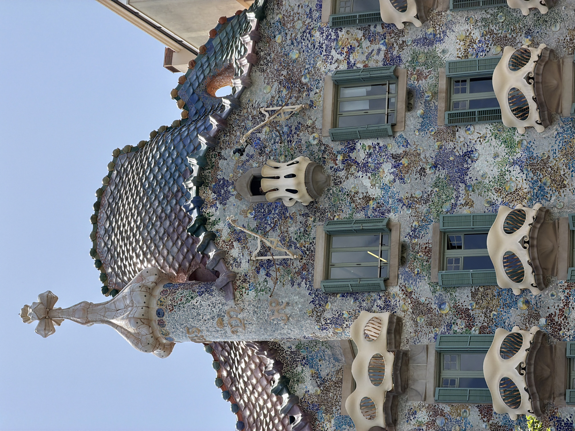 This is a close-up, low-angle photograph of the exterior of Casa Batlló in Barcelona, Spain, a building designed by Antoni Gaudí. The image is oriented vertically, with the sky taking up the upper left portion.
The building's facade is adorned with a mosaic of colorful tiles in shades of blue, green, purple, and brown, interspersed with glass elements that create a shimmering effect. The architectural style is highly organic and fantastical. On the left side of the frame, a distinctive roof feature resembling a dragon's spine, covered in iridescent scales, dominates the image. Further down, a pointed, cross-like finial extends outwards.
Numerous windows are visible, many with green shutters. Above some windows, the facade features unique, bone-like organic sculptures, reminiscent of masks or skeletal structures, with striped openings. These decorative elements and the overall mosaic pattern are characteristic of Gaudí's modernist style.
The scene appears to be set on a bright, clear day, indicated by the deep blue of the sky and the strong lighting. The sun illuminates the intricate details of the building, highlighting the textures and colors of the tiles and sculptures. There is no visible text in the image. The focus is entirely on the unique and artistic architecture of the building.