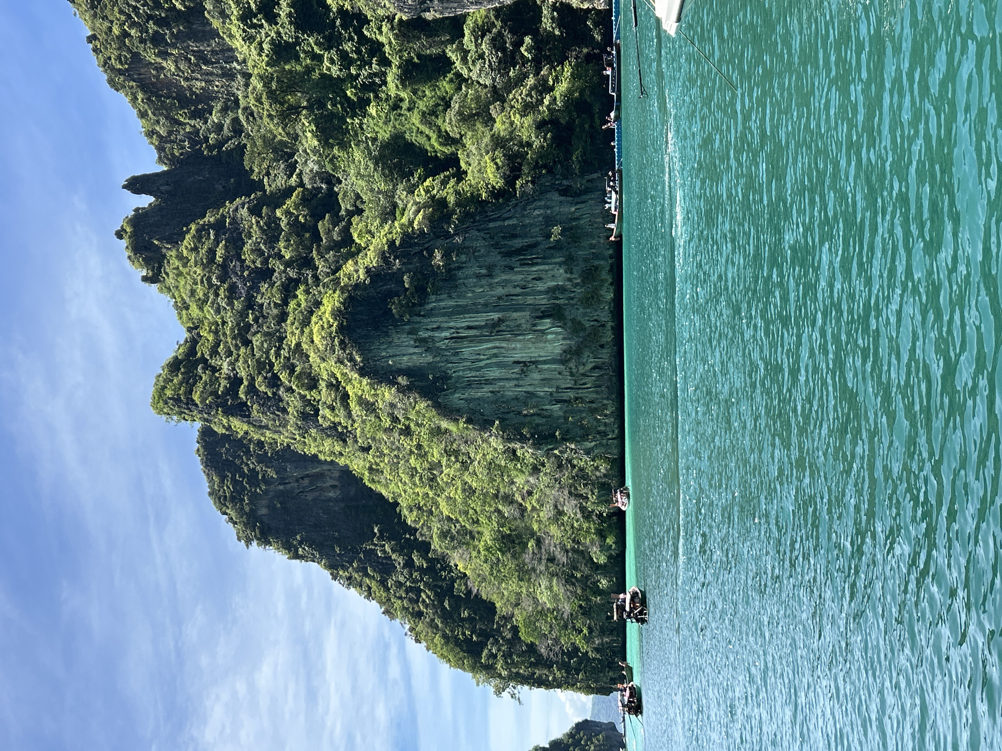 This image captures a tropical seascape in Ao Nang, Thailand, featuring towering limestone karsts emerging from vibrant turquoise waters under a clear blue sky with sparse white clouds. The karsts are heavily vegetated with dense, bright green foliage, exhibiting steep, rugged cliff faces and prominent vertical striations on one large rock formation. Several traditional Thai longtail boats are dispersed across the water in the midground, each carrying small groups of individuals. The water's surface displays small ripples and reflects sunlight, transitioning from a deeper emerald shade in the foreground to a lighter turquoise closer to the islands. A portion of a white boat's hull and mooring line is visible in the bottom right corner, indicating the viewer's perspective from a boat.