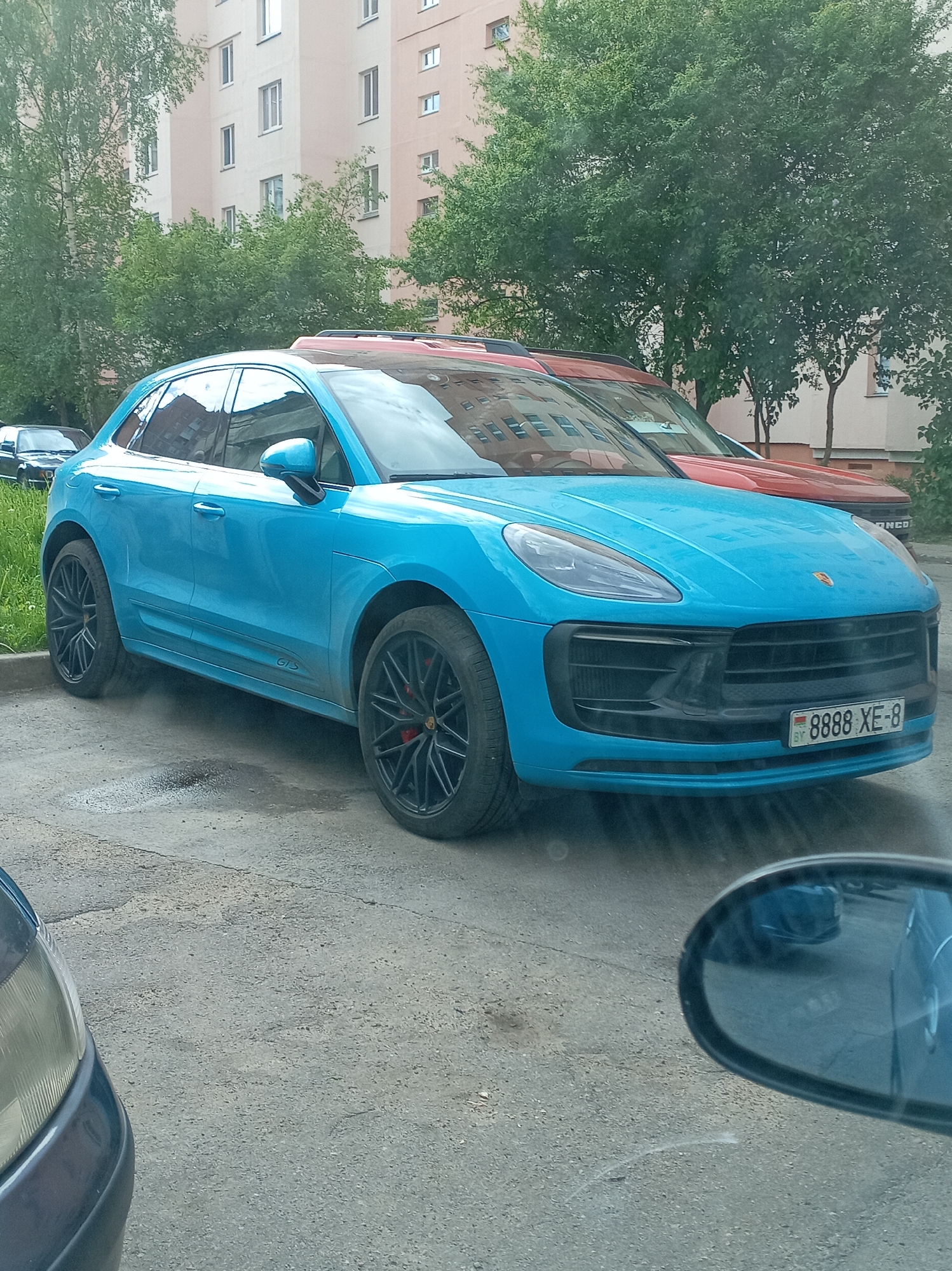A light blue Porsche Macan GTS is parked on an asphalt surface in an urban environment. The car has black rims with red brake calipers and a Belarusian license plate reading "8888 XE-8." Behind the Porsche, a red SUV is partially visible. In the background, there's a light-colored apartment building and several green trees. A portion of another blue car and its side mirror are visible in the foreground. The location is in the city of Unknown, Unknown.