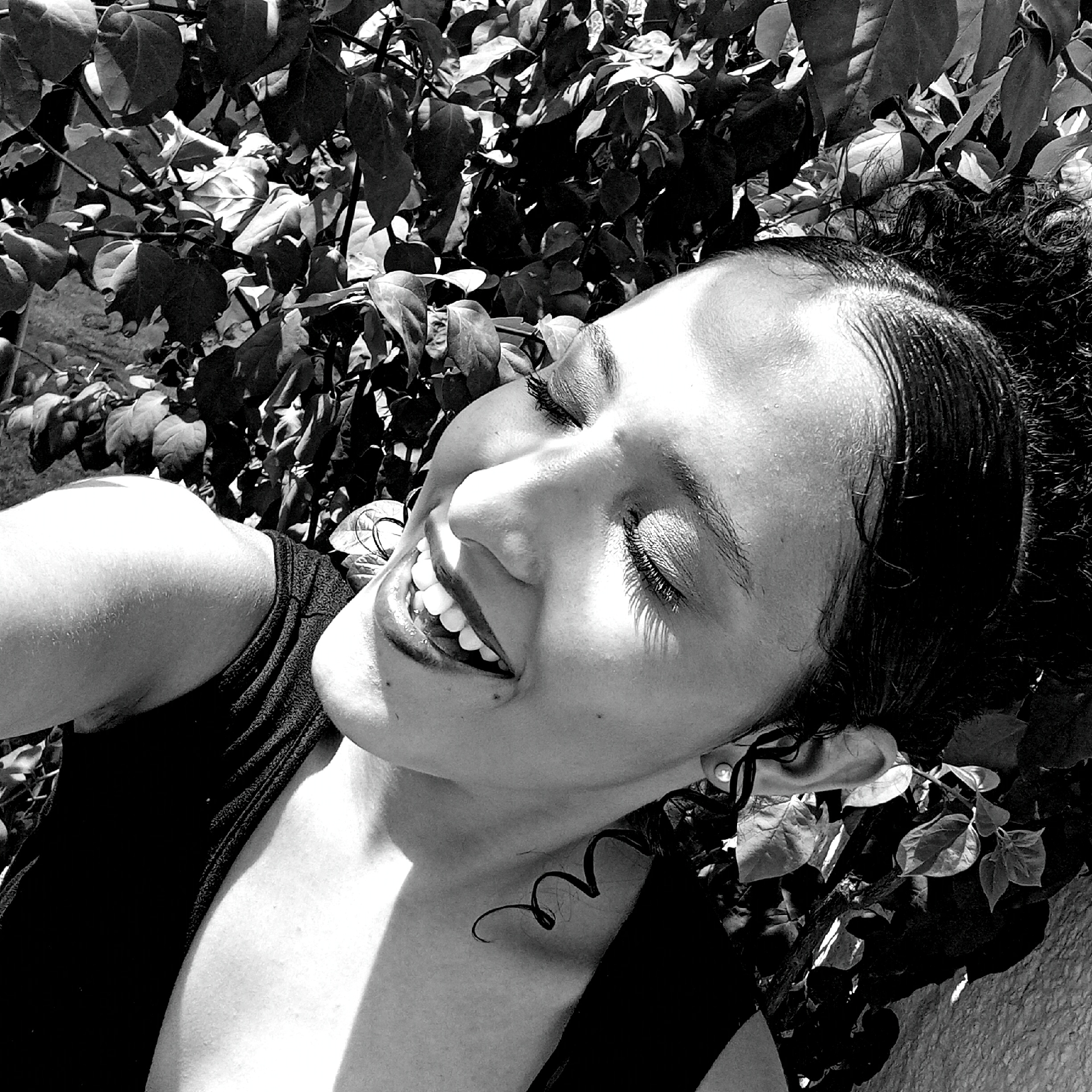 This black and white, close-up photograph captures a woman lying outdoors, her head nestled amidst a dense bed of foliage, likely in Parroquia Ocumare, Venezuela. She is seen from an elevated perspective, looking upward with her eyes closed and a broad, genuine smile revealing her teeth, conveying a feeling of joy, laughter, or deep contentment. Strong, direct sunlight casts bright highlights across her face and arm, creating distinct shadows that suggest a clear, sunny daytime environment. Her dark, curly hair is visible, with some strands appearing slicked back from her forehead. A prominent, well-defined curl rests near her neck. She is wearing a dark, possibly sleeveless, top. An earring adorns her visible ear. The background is dominated by the textured, heavily shadowed leaves and branches of a bush or tree, contributing to the natural setting. No text is visible in the image.